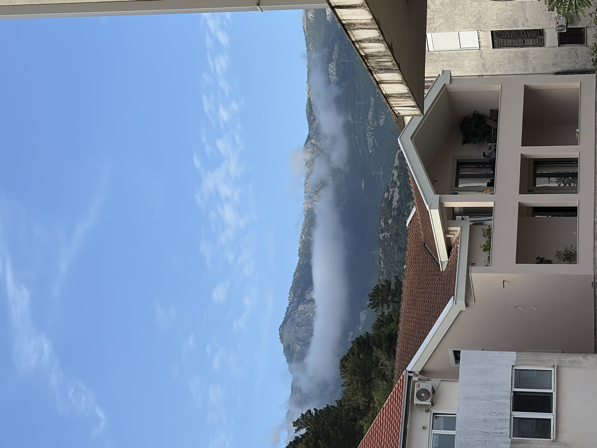 The image shows a view from a building in what is likely Budva, Montenegro, capturing a slice of the city's built environment alongside a mountainous landscape.
The sky is predominantly blue with sparse cirrus clouds. A mountain range is visible, partially obscured by fog or low-lying clouds clinging to its slopes. The mountain is covered in vegetation.
In the foreground, the architecture of Budva is evident. Buildings with red-tiled roofs and neutral-colored facades are prominent. A multi-story building with open balconies and rectangular openings is also visible. An air conditioning unit is mounted on one building. The buildings have windows of varying sizes. The angle of the shot suggests that the image was taken from within another building or from a raised vantage point.