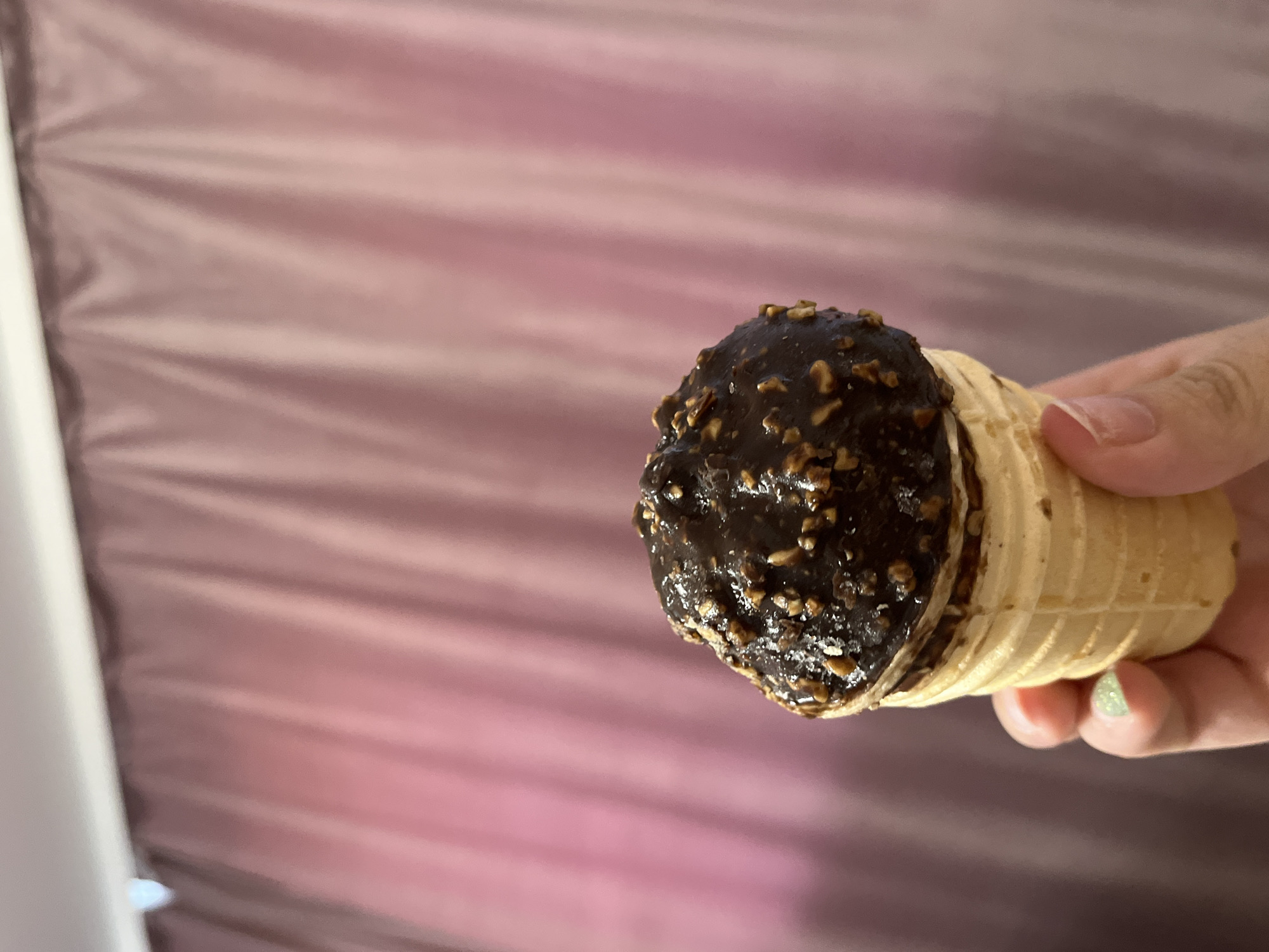 A hand holds a chocolate-covered ice cream cone with chopped nuts against a backdrop of pink, draped curtains. The ice cream is domed and coated in dark chocolate with visible pieces of nuts. The cone appears to be a light brown waffle cone. The background is out of focus, showing the texture and color of the curtains. The lighting suggests an indoor setting. There is no indication of the location being Krasnyye Barrikady, Russia.