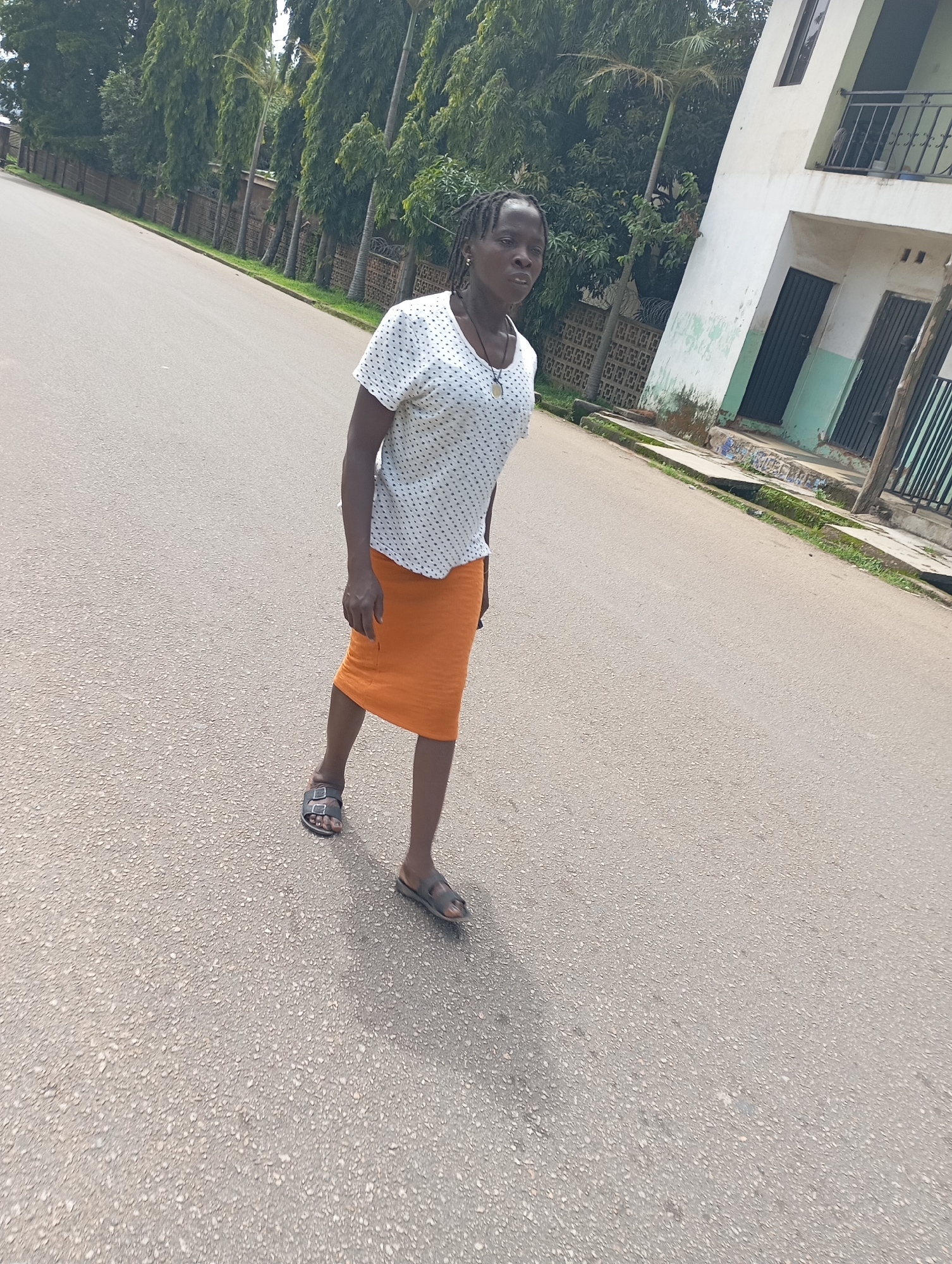 An African woman walks down a paved road in Jos, Nigeria. She is wearing a white polka dot shirt and an orange skirt, with dark sandals. The road is lined with trees and a fence on the left, and a light-colored building with black-barred windows and doors on the right. The sky is overcast.