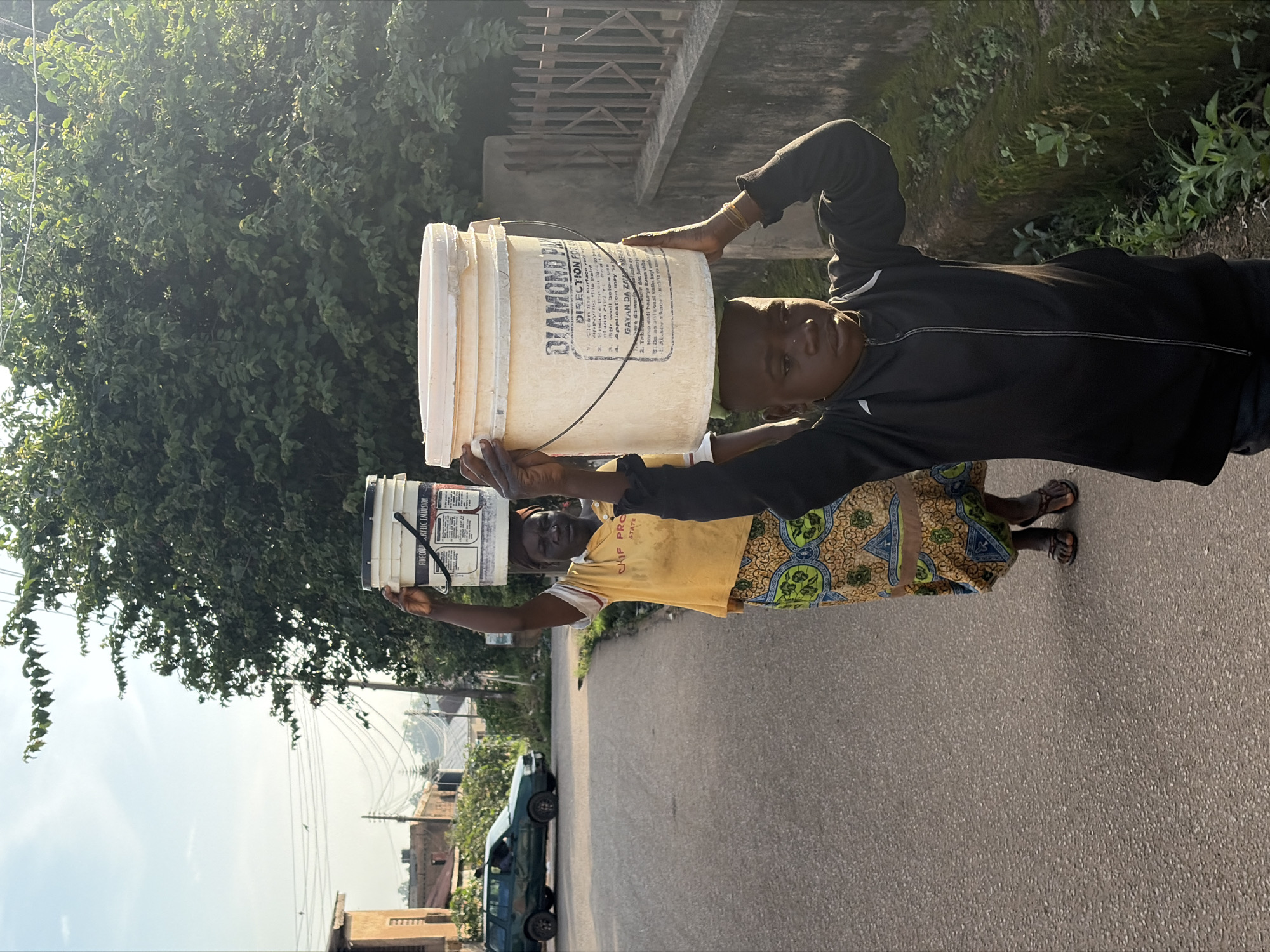 In the city of Jos, Nigeria, a young boy and a woman are captured from a side profile, walking down a paved road. The boy, positioned in the foreground, carries a large, light-colored bucket filled with a white substance on his head. He is dressed in a black zippered jacket over dark pants. Behind him, the woman, wearing a yellow t-shirt with an orange and green patterned skirt, also balances a white bucket on her head. The road they are walking on is a two-lane asphalt surface with a worn appearance. To the left of the road, a green SUV is parked, and beyond that, a two-story building with an orange facade is visible. Further in the background, houses and trees line the street, with power lines stretching across the sky. The right side of the image features a low concrete wall covered in moss and overgrown with plants. The lighting suggests it is either early morning or late afternoon, casting long shadows.