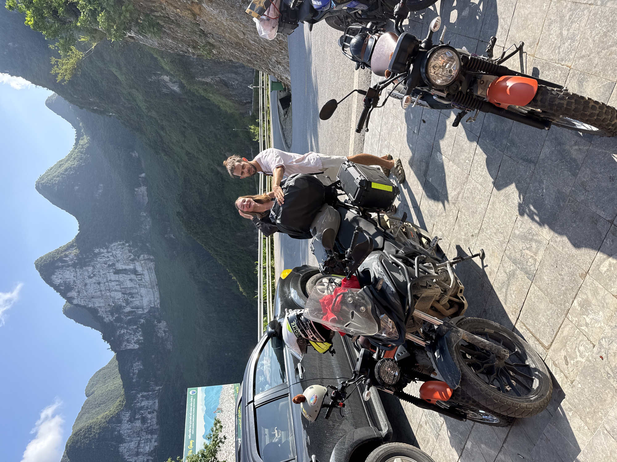 The image shows a couple posing next to two motorcycles and a dark-colored car on a paved surface in the Đồng Văn Commune, Vietnam. The landscape features tall, steep, green-covered mountains under a clear, blue sky. The motorcycles appear to be adventure bikes, loaded with luggage. The couple is close to one of the bikes, with the man standing behind the woman. A sign is visible in the lower left corner.