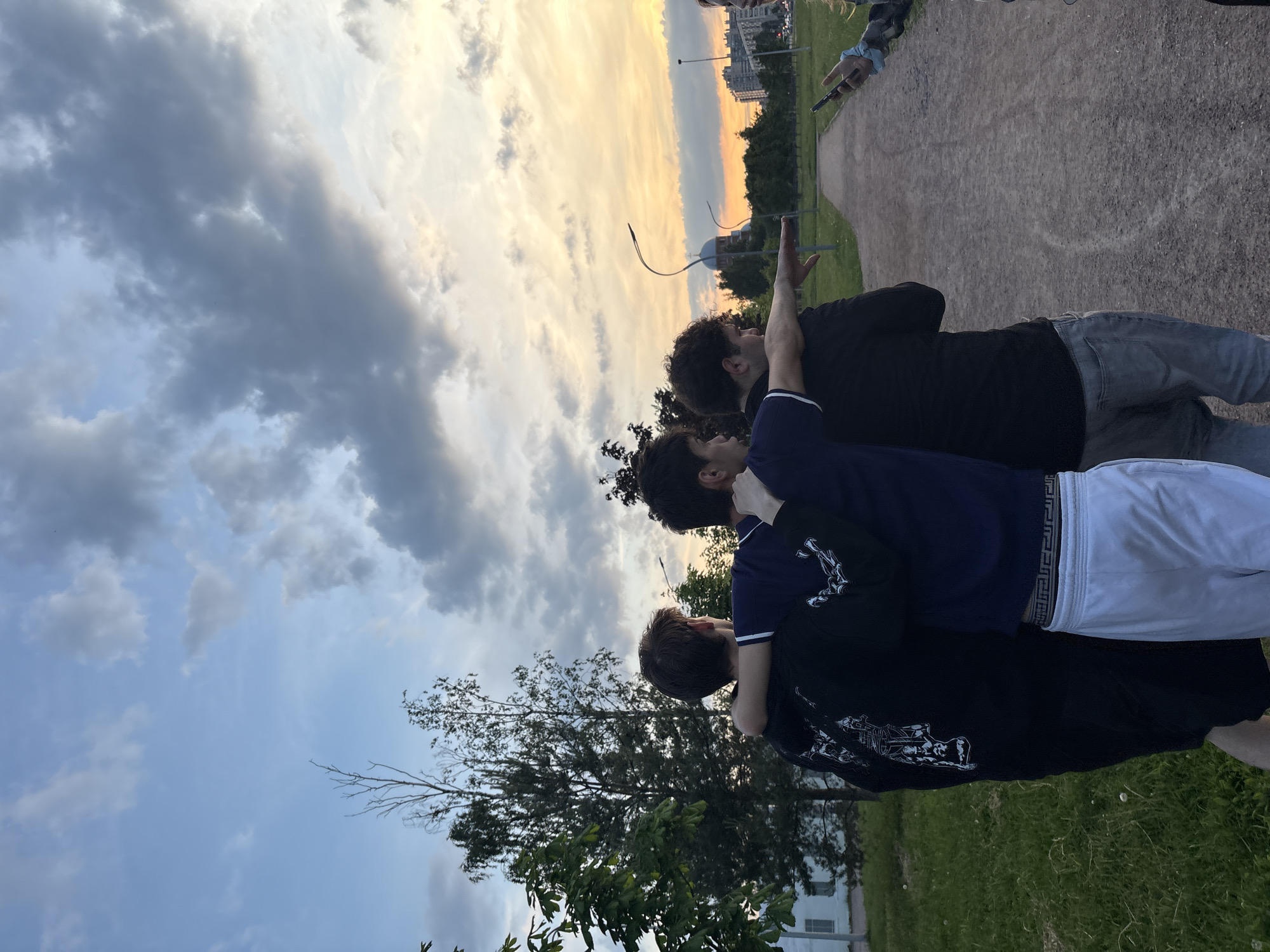 In the city of Sinyavino, Russia, at dusk, three young men stand with their backs to the camera, their arms around each other's shoulders, facing a park-like area with a gravel path. The sky above is a mix of blue and grey clouds, with hues of orange and yellow from the setting sun visible on the horizon. Trees line the park, and in the distance, there are apartment buildings and a domed structure. One of the men is pointing towards the distance, while another is holding a smartphone. The men are dressed in casual clothing: a black hoodie, a blue t-shirt, and a black t-shirt with grey jeans. The environment suggests a warm evening.