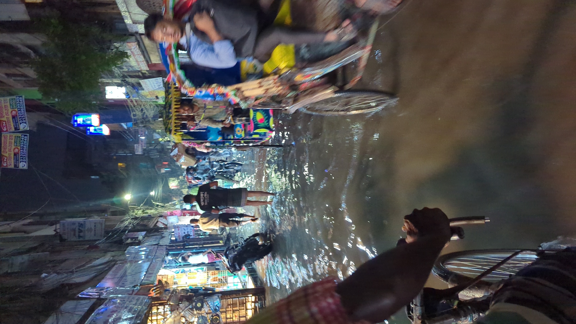 The image depicts a flooded street in Dhaka, Bangladesh, at night. People are wading through the water, and rickshaws are being used to navigate the flooded area. The water appears murky and reflects the lights from nearby buildings. Numerous shops and street signs are visible along the sides of the street. The foreground shows the hands of a person holding onto bicycle handles, presumably riding or pushing it through the water.