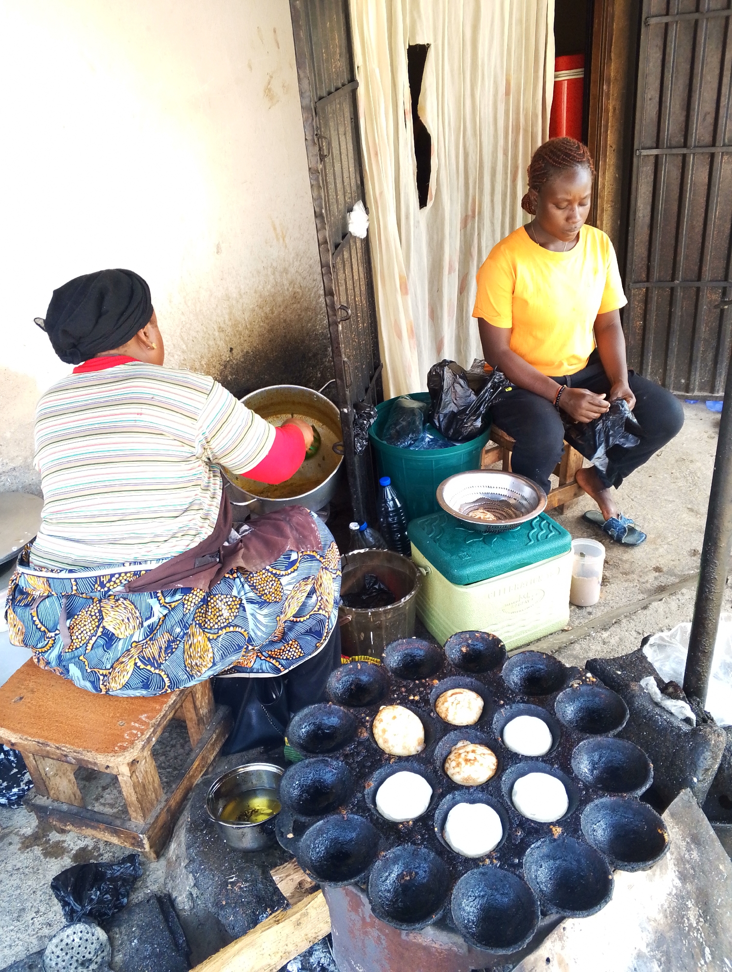 In Jos, Nigeria, two women are engaged in food preparation outdoors, likely at a street stall. The woman on the left, wearing a striped shirt and a colorful patterned skirt, is seated and stirring a large pot of food. The woman on the right, in an orange t-shirt and black pants, is sitting and appears to be holding a plastic bag, with a green cooler and other items around her. In the foreground, a dark, circular cooking apparatus with multiple indentations is visible, containing several round, white food items that are in various stages of cooking. A small metal pot with yellow liquid, presumably oil, is also nearby. The environment appears to be a somewhat rudimentary outdoor setting, with a wall behind the women and a metal gate or fence to the right. The ground is concrete or packed earth.