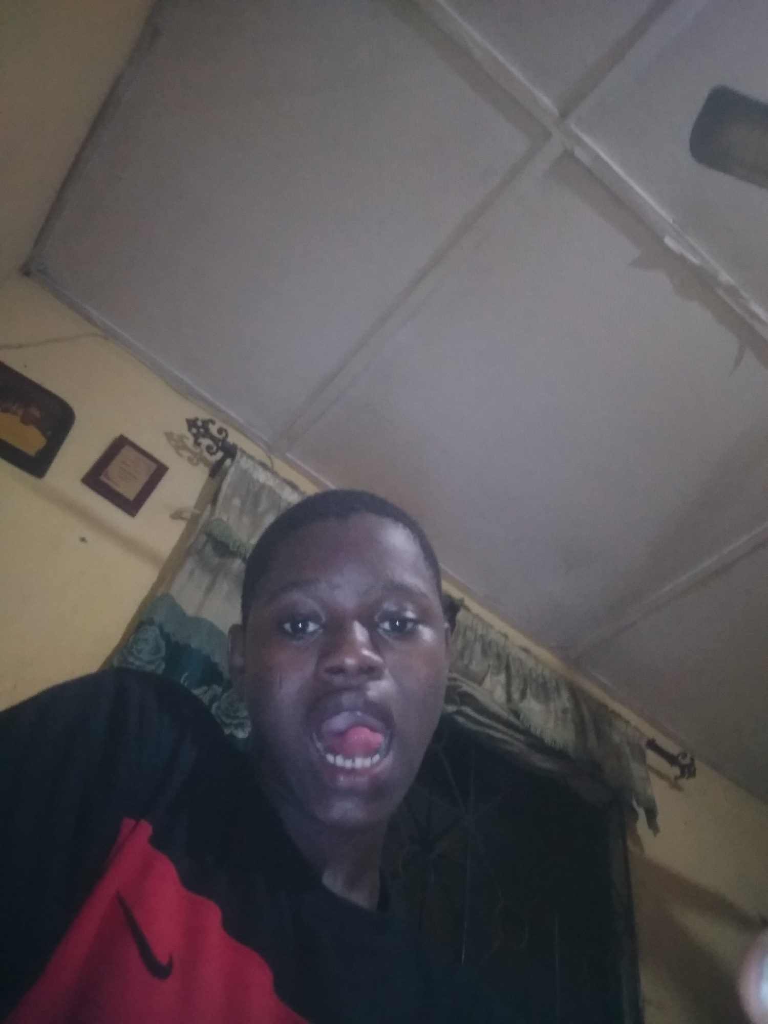A young Black male is in a room in Unknown, Unknown. He is wearing a black and red t-shirt and has his mouth open. The room has a white ceiling, yellow walls, a window with curtains, and some framed pictures on the wall.