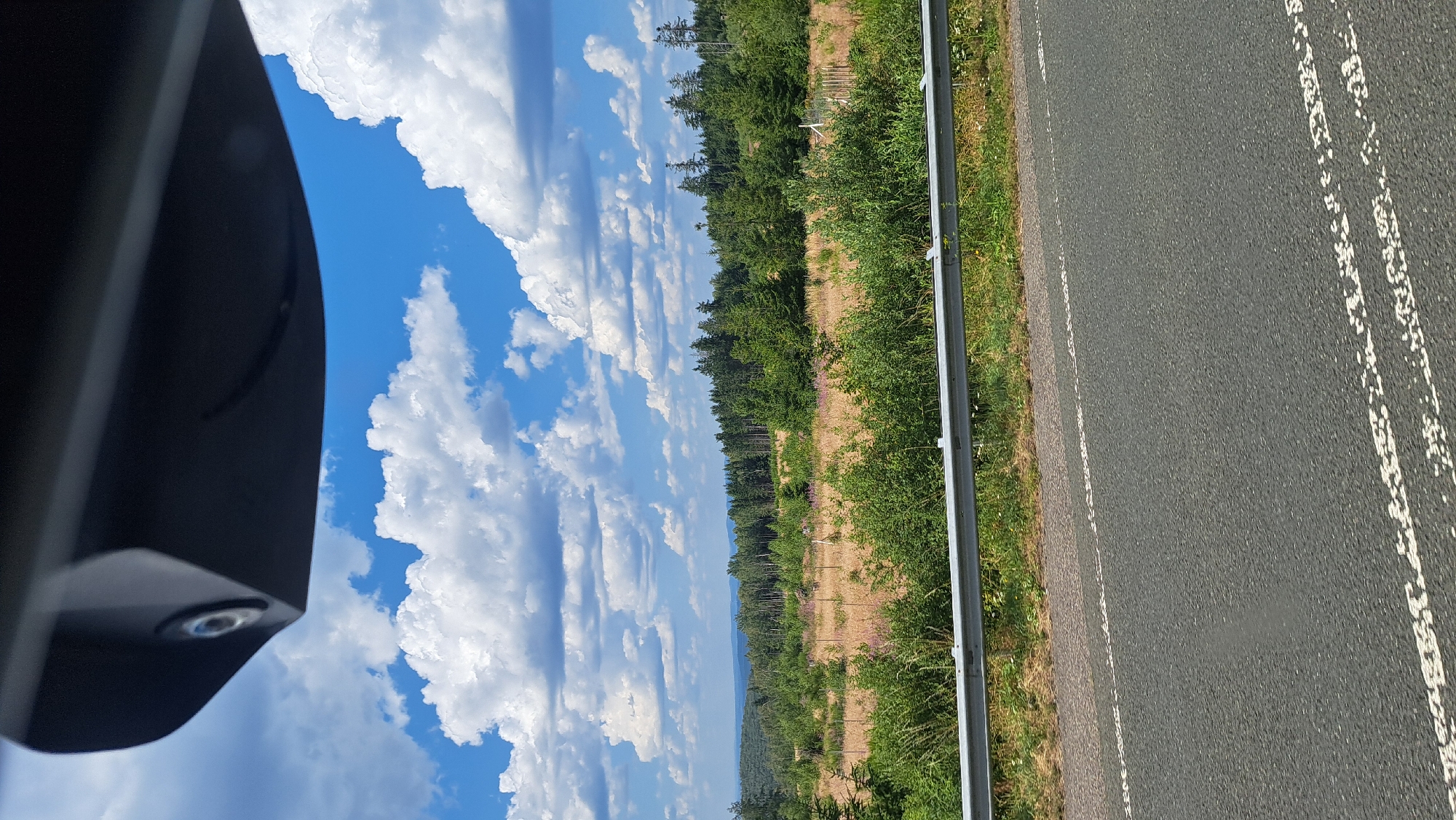 The image shows a landscape scene from a vehicle's perspective, looking out towards a forest and fields under a blue sky with white clouds. In the foreground, the asphalt road with white lane markings is visible, bordered by a metal guardrail and grassy embankment. The middle ground features a dry, grassy field with some scattered pink wildflowers, transitioning into a dense forest of evergreen trees. The background displays rolling hills and more distant trees. A dark, curved object, likely a vehicle's rearview mirror or a similar component, is positioned at the top of the frame. No people or animals are discernible in the image. The scene suggests a rural or semi-rural location. Based on the query, the location is identified as Burbach, Germany.