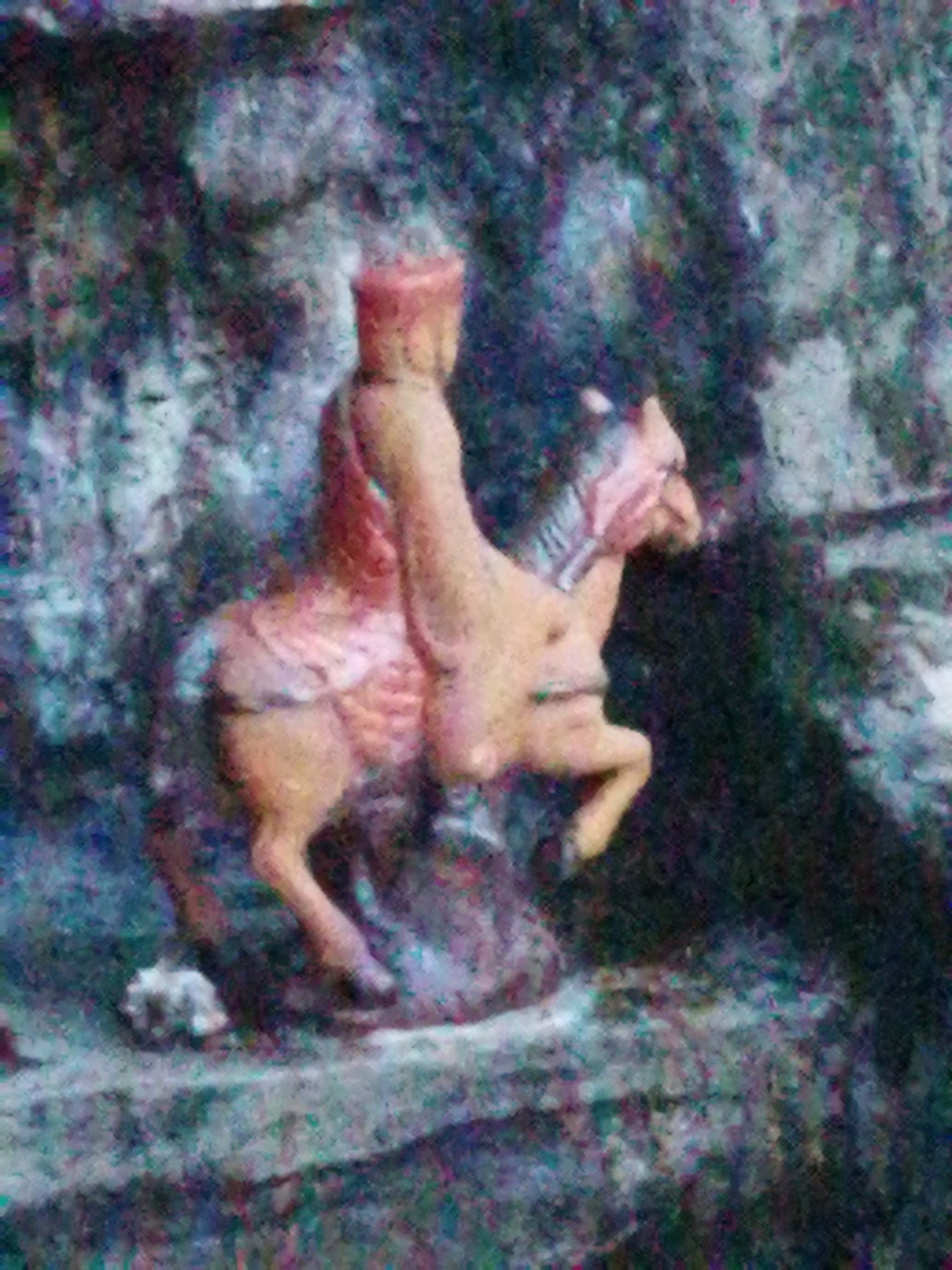 A statue depicts a man riding a horse. The man wears a red hat and a long, tan coat. The horse is tan and is rearing up on its hind legs. The statue is located outdoors and appears to be part of a larger structure, possibly a temple or shrine. The background is a mix of rock and vegetation. This could be located in Da Lat, Vietnam.
