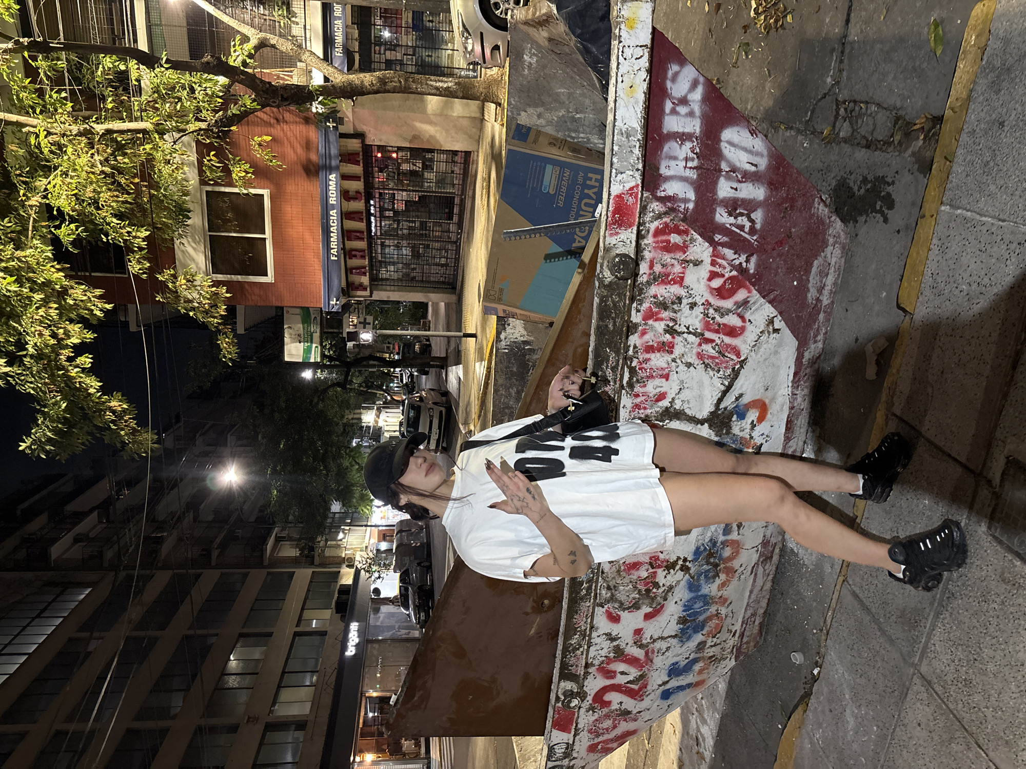 A young woman stands inside a metal dumpster on a city street. She wears a white oversized t-shirt, black sneakers, and a black baseball cap. She has tattoos on her arms and is holding a small black purse or bag. The dumpster is filled with trash and covered in graffiti. The location is in Buenos Aires, Argentina, indicated by the "Farmacia Roma" sign visible in the background, and the "brigatti" sign on a building. It is nighttime, illuminated by streetlights. Buildings line the street.