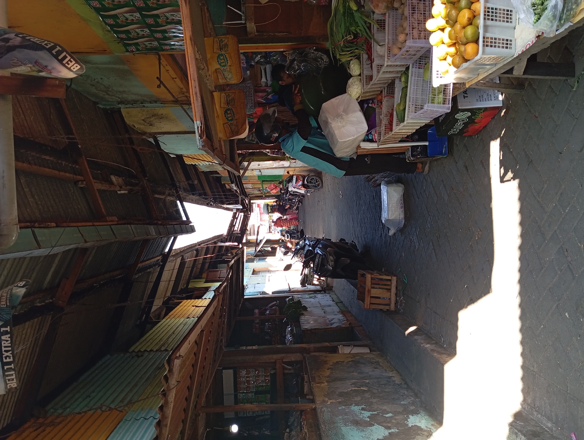 The image depicts a narrow outdoor market alley in Kepulungan, Indonesia, under a corrugated tin roof structure. The alleyway is paved with interlocking bricks and is lined with market stalls. On the left, a stall displays various items, including what appears to be packaged goods. On the right, a vendor, wearing a blue shirt and a helmet, is interacting with a customer. The vendor is handing over a white package, while the customer is partially obscured. In front of the vendor's stall, there are several crates filled with fresh produce, including cucumbers, cabbage, chilies, and citrus fruits. Several motorcycles are parked along the alley, and a woman in a pink headscarf is visible in the background. The sunlight casts sharp shadows across the ground, indicating it is daytime. The market appears to be active, with goods displayed and people engaged in transactions.