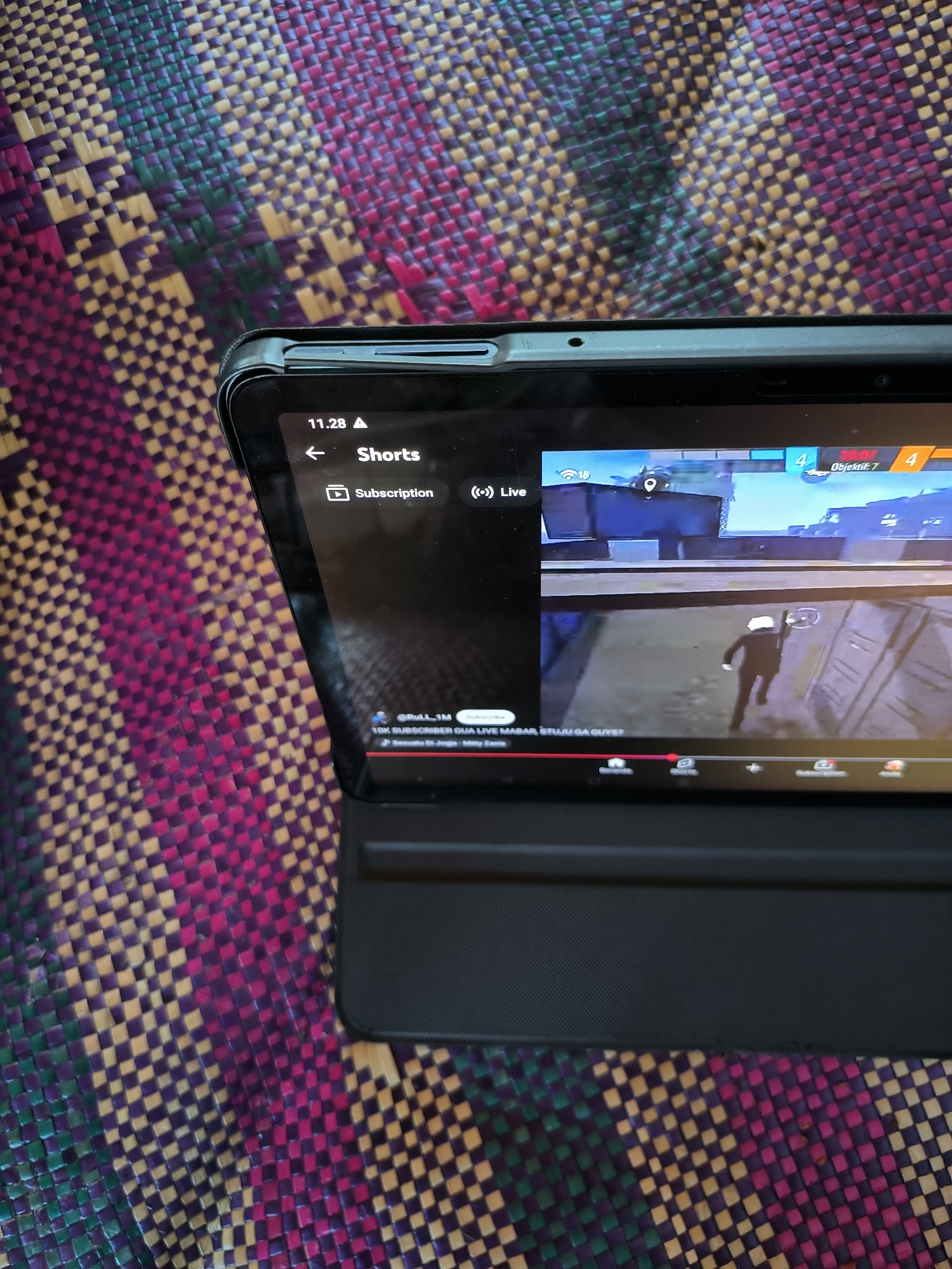 A tablet displays a live stream of a game similar to Free Fire on a woven mat. The screen shows a character in a black suit and white mask in a white building. On the top of the screen are the words "Shorts," "Subscription," and "Live," along with game statistics. The tablet is on a mat woven with colors purple, green, and yellow. The OCR indicates the location of Merbopuntong, Indonesia.