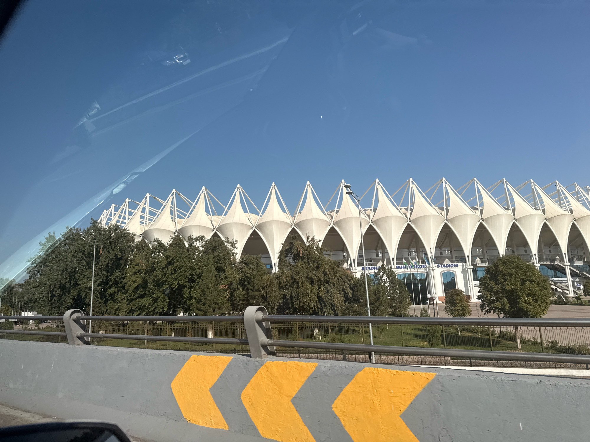 The image, captured from a vehicle in Tashkent, Uzbekistan, shows a view of the Humo Arena, a modern indoor sports and concert venue. The arena's distinctive white, tent-like roof structure is prominently displayed against a clear blue sky. In the foreground, a grey concrete barrier with bright yellow chevron markings is visible, suggesting a roadway or overpass. Behind the barrier, a metal railing separates the roadway from a landscaped area with lush green trees. Flags are flying in front of the arena's entrance, and signage indicates it is a stadium. Reflections of the sky and possibly the vehicle's interior are visible on the arena's windows. The overall scene depicts a sunny day in a vibrant urban environment, highlighting a key piece of Tashkent's modern infrastructure.
