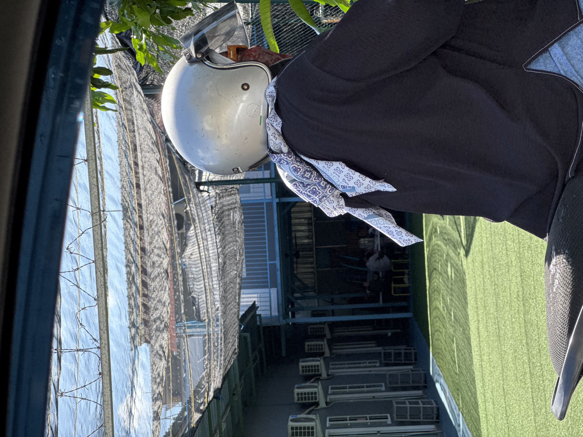 A medium shot, taken from a slight angle, shows a person wearing a silver motorcycle helmet and a black jacket. A blue and white patterned scarf is tied around the helmet. The person is seated on a motorcycle. The background features a building with blue metal frames and rows of air conditioning units along the side. A black mesh-like covering is visible on the upper left side of the frame, partially obscuring the sky. A patch of green artificial turf is also visible. The location is in Kuta Selatan, Indonesia.