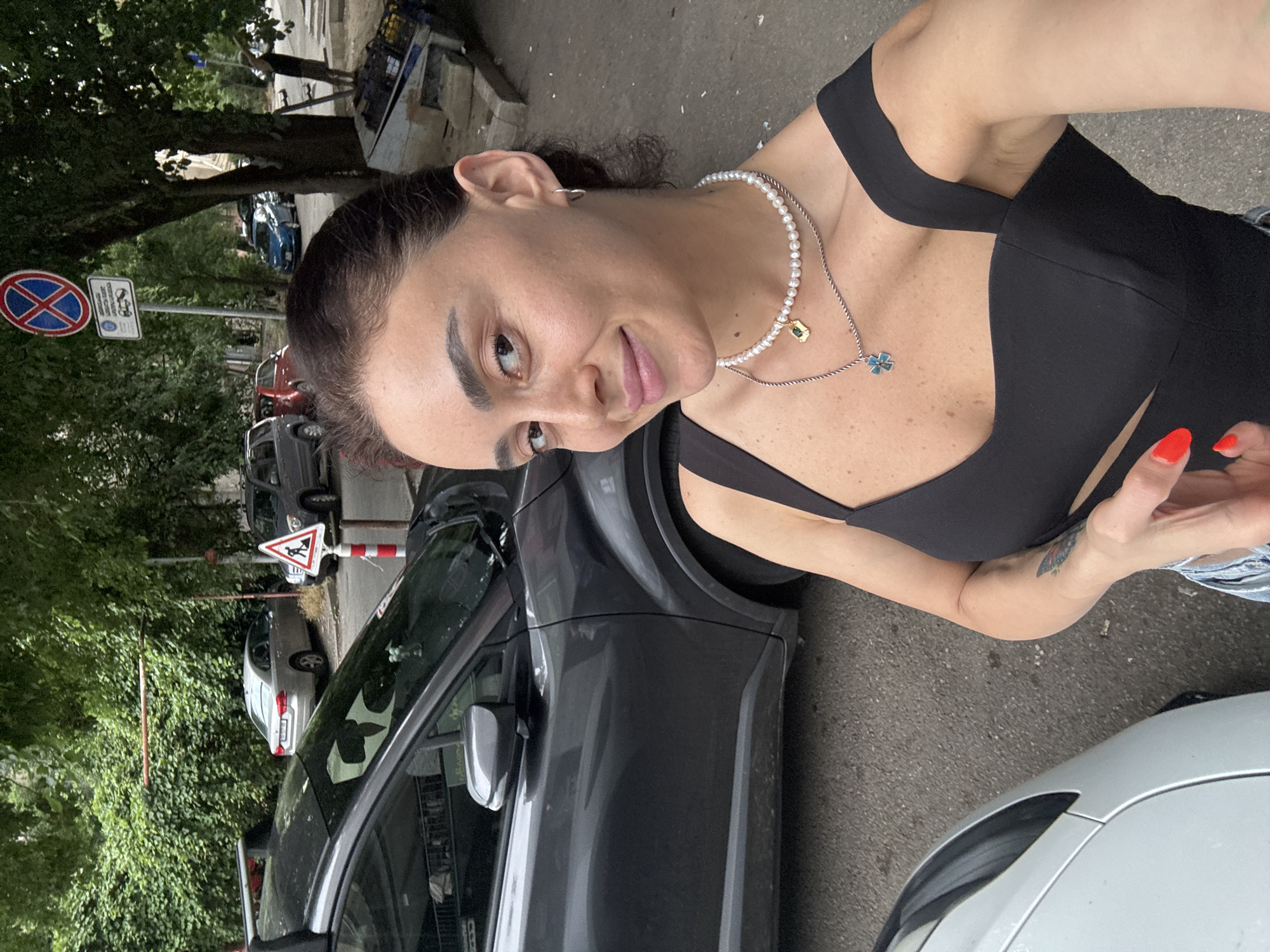The media file shows a woman standing on a street in Tbilisi, Georgia. She is wearing a black top and a pearl necklace. She has bright red nail polish on her fingernails. Cars are parked on the street behind her, and there are trees and buildings in the background. There are also traffic signs visible, including a "no parking" sign and a sign indicating a pedestrian crossing. The lighting suggests it is daytime. The woman appears to be looking at the camera with a slight smile.