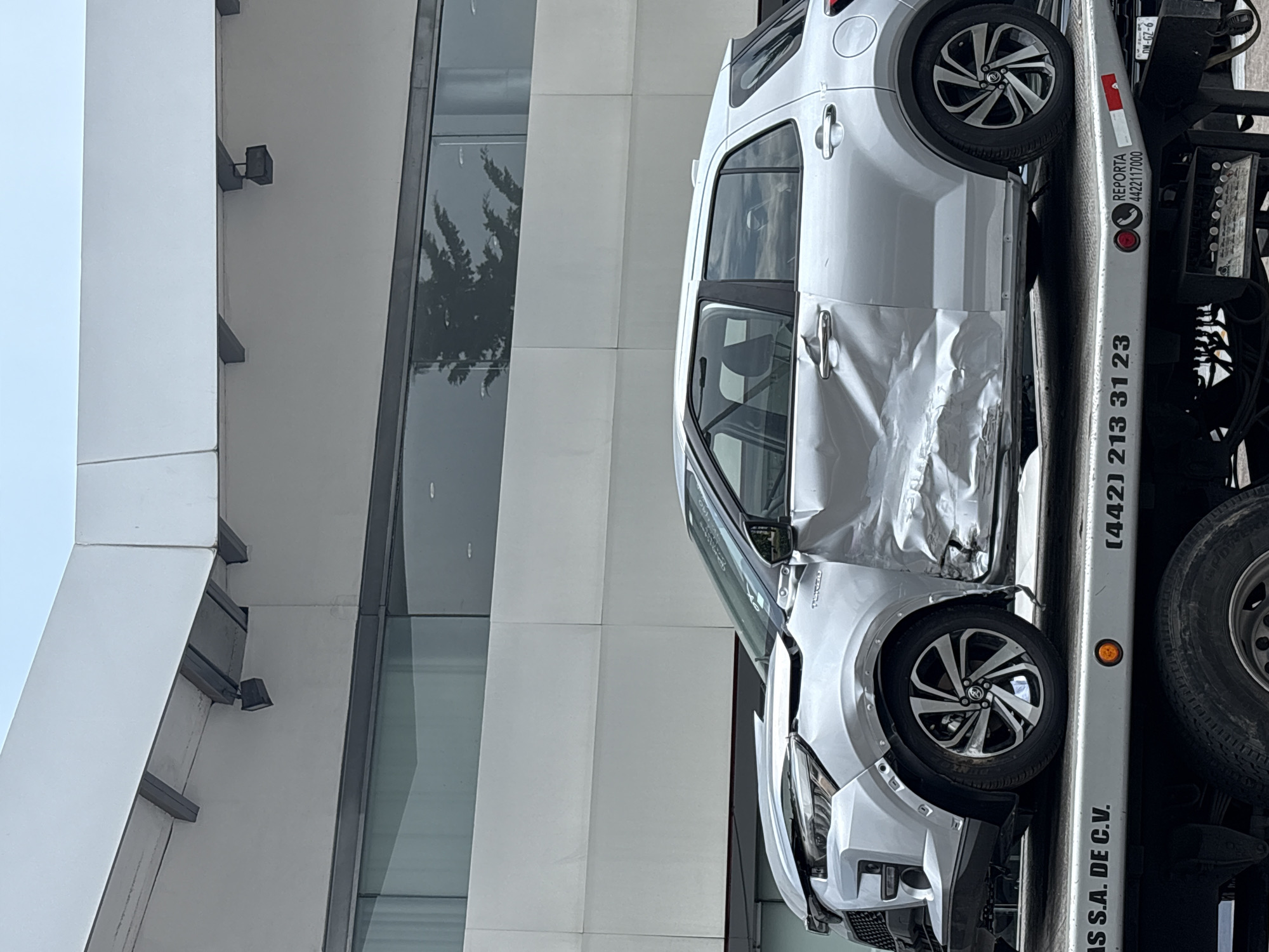 The image, initially rotated 90 degrees counter-clockwise, depicts a silver compact car, likely a Toyota Yaris Cross, with significant damage being transported on a flatbed tow truck. The scene unfolds in an outdoor setting during daytime, adjacent to a modern building.
The car shows severe impact damage along its entire driver's side, with both the front and rear doors visibly crumpled and distorted. The front fender and headlight on that side are also damaged. The distinctive two-tone alloy wheels appear structurally intact.
Only a portion of the flatbed tow truck is visible, displaying its light grey surface, control console, and a spare tire. The building in the background features a clean, modern design with expansive reflective glass windows that mirror surrounding trees, and light-colored, possibly white or light grey, paneling. The bright, even lighting suggests a clear day.
Several pieces of text are visible on the side of the tow truck: "AS S.A. DE C.V.", along with two phone numbers "(442) 213 31 23" and "REPORTA 4422117000". Other smaller text includes "SODWER" (partially visible on the spare tire) and "0.0", "9-29-NO" which might be part of vehicle identification or a license plate. The Spanish text and the (442) area code, which belongs to Querétaro, are consistent with the provided location context of Mexico, where the vehicle is being transported following an apparent accident.
