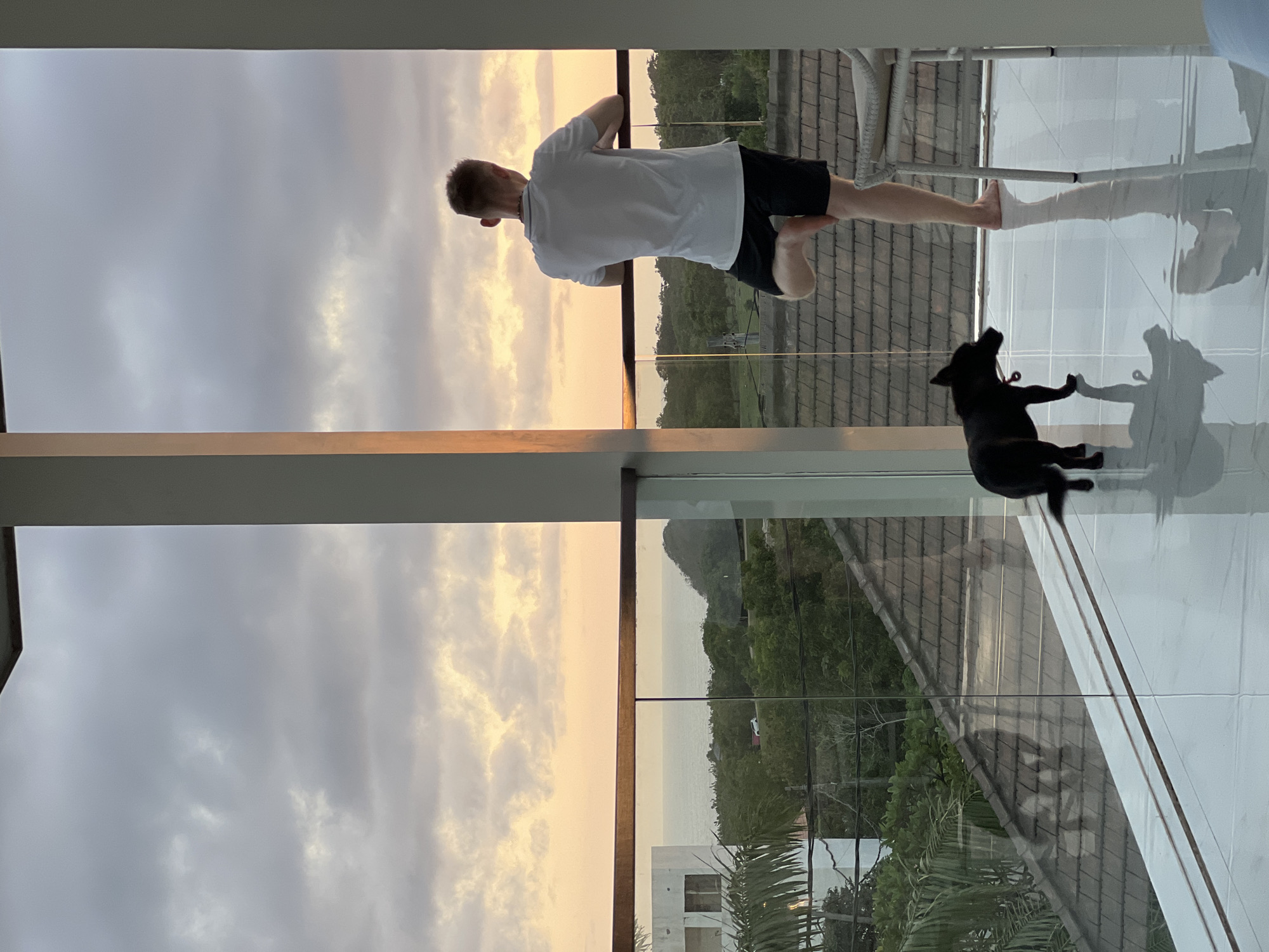 A landscape view of an individual standing on a porch with a black dog laying down. The individual is white, has short brown hair and is wearing a white t-shirt and black shorts. The dog is black and appears to be resting on the white tiled porch. The porch overlooks an outside view of trees, and a body of water with building structure in the foreground. The sky is overcast, and the time of day appears to be either sunrise or sunset. The location is Unknown, Unknown.
