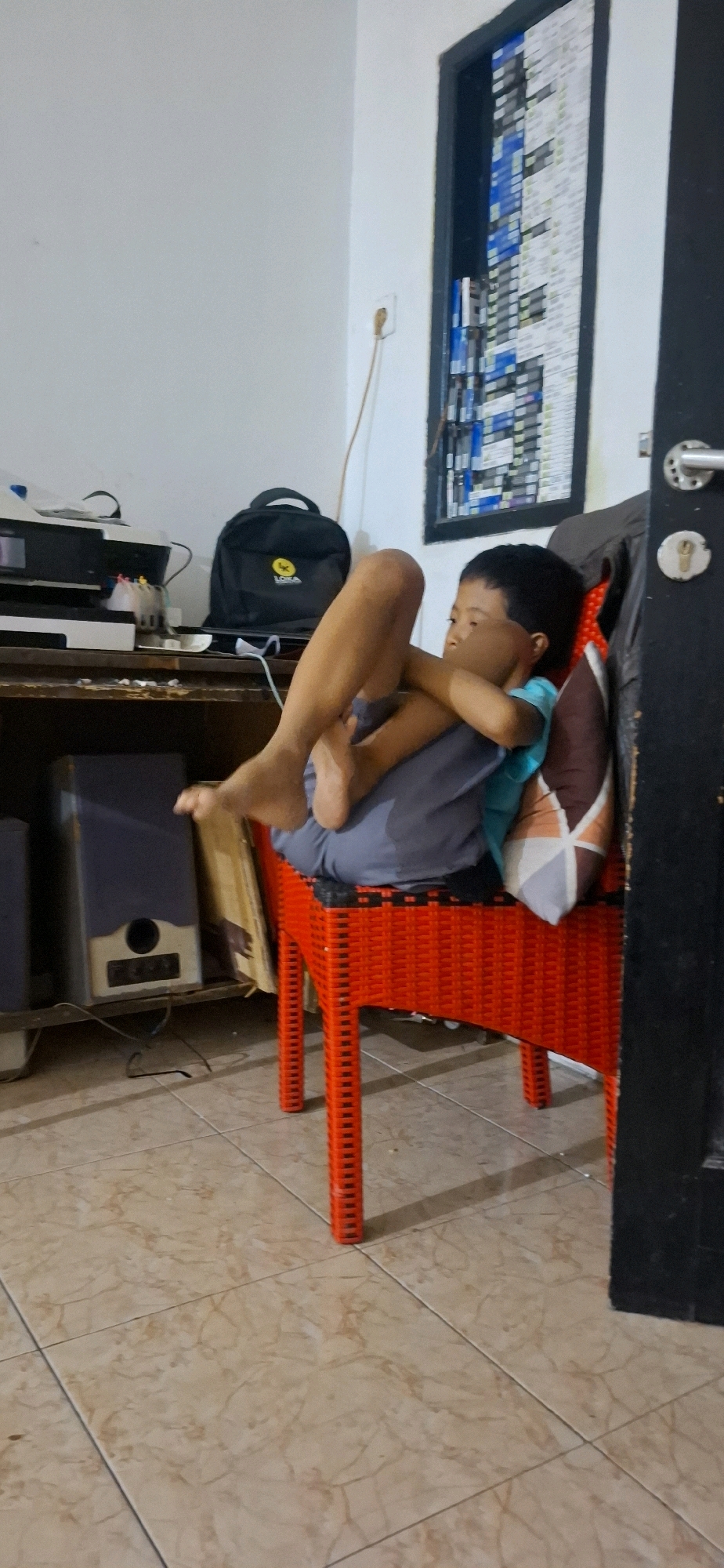 A young boy is sitting on a red plastic chair with his legs crossed and his arms wrapped around his knees. He is wearing a light blue t-shirt and dark grey pants. The boy is looking to his left, away from the camera. The chair is positioned in front of a wooden desk with various items on it, including a printer, a black backpack, and some papers. To the left of the desk are two large speakers. The room has a tiled floor and a white wall. A door with a black frame is visible on the right side of the image, with a keyhole and a doorknob. The door is decorated with a grid of small, rectangular items, possibly files or cards. The scene appears to be an indoor residential or office space in Ciharashas, Indonesia.