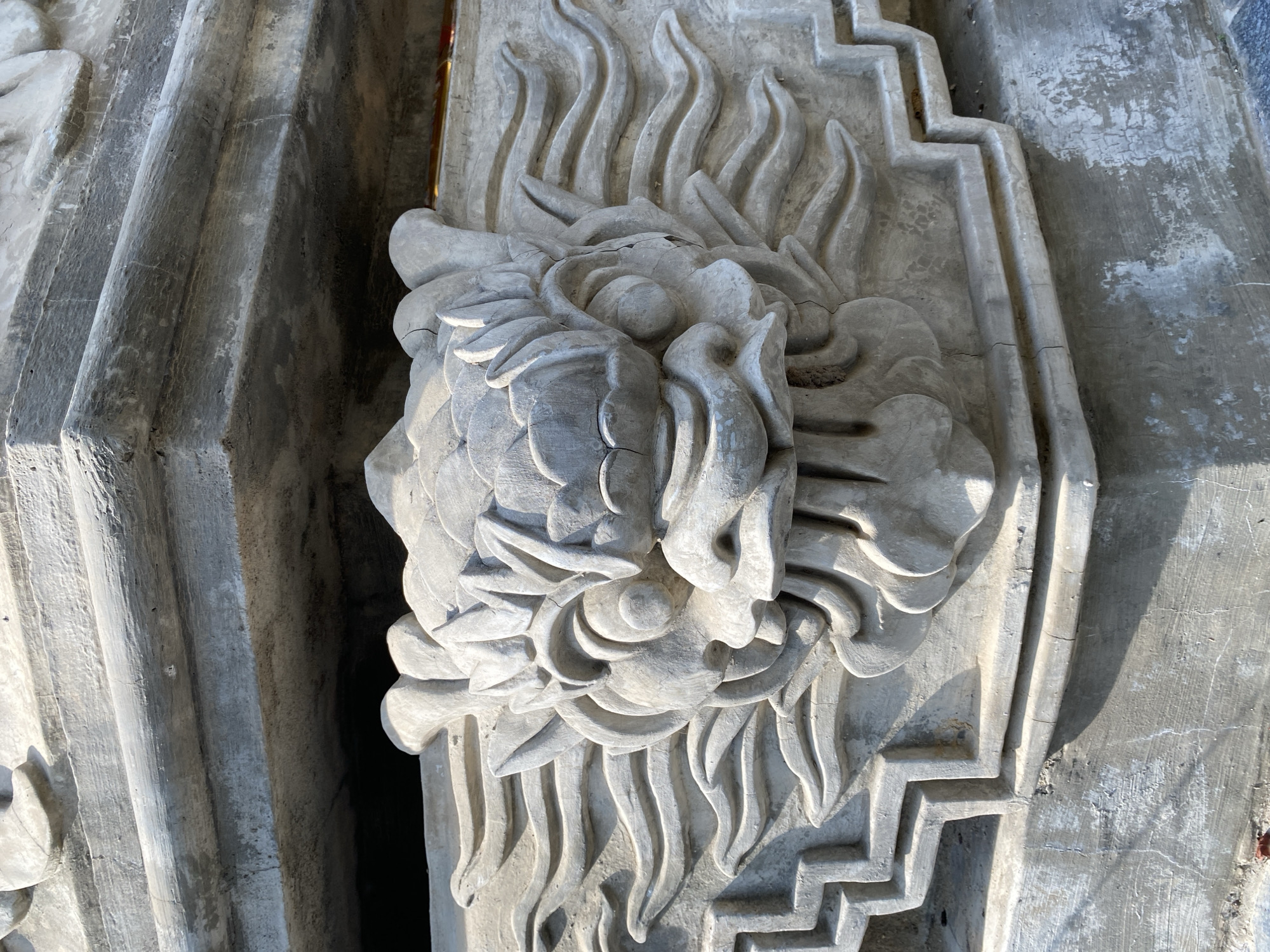 The image shows a close-up, detailed view of a weathered, grey stone carving of a dragon's head. The carving is ornate, with intricate details of scales, horns, and a fierce expression. It appears to be part of a larger architectural element, possibly a building or a monument. The stone shows signs of age and exposure to the elements, with some cracks and chipping visible. The lighting suggests it is daytime, with sunlight casting shadows on the carving. The surrounding structure is also made of stone and has a layered, stepped design. The dragon carving is positioned centrally and prominently, indicating its significance. The overall impression is one of ancient artistry and enduring craftsmanship. The location is in Tay Nha Trang Ward, Vietnam.