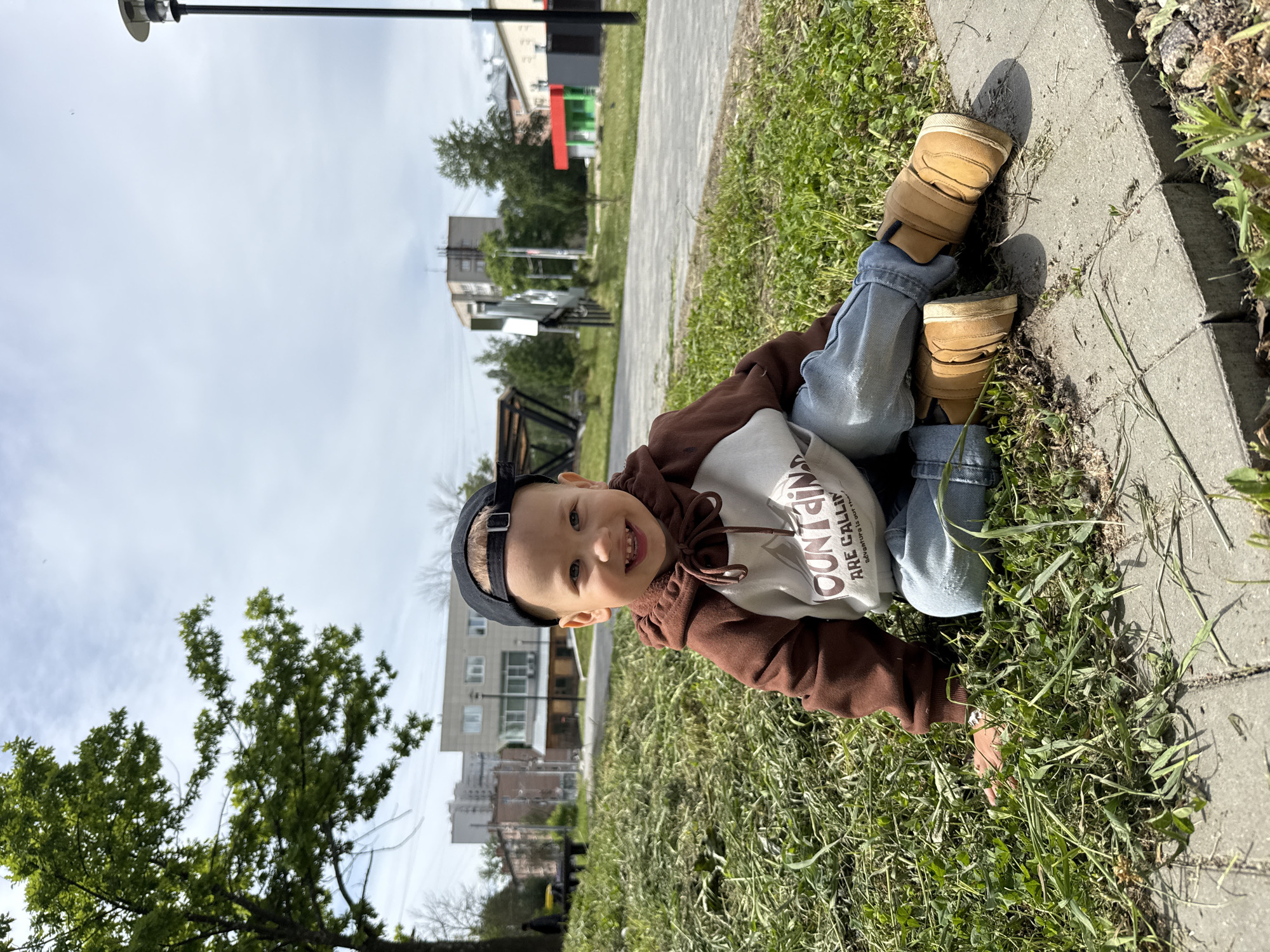 A young child wearing jeans, brown boots, and a brown and white hooded sweatshirt with a backwards black baseball cap is lying on the grass in Rusanovo, Russia. The child is smiling at the camera, while buildings and a street lamp are in the background. A paved pathway runs along the edge of the grass where the child is lying.