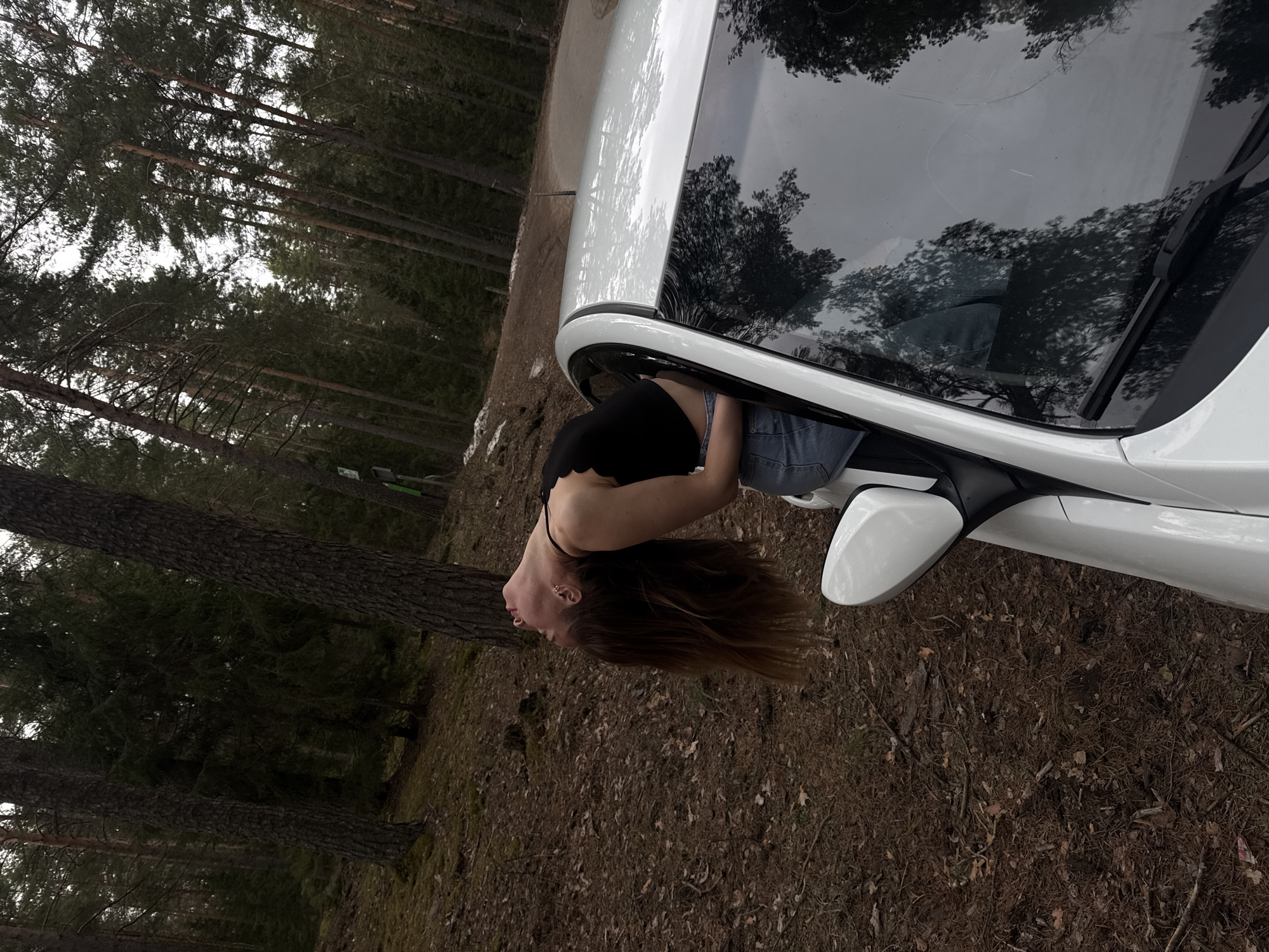 A young woman is captured in an unusual pose, leaning out of the passenger window of a white car in a forest setting in Chaikovsky, Russia. Her upper body is inverted, with her head hanging downwards towards the ground. She wears a black, scalloped-edge crop top and denim bottoms, possibly shorts or jeans. Her long, dark brown hair cascades freely towards the forest floor. An earring is visible in her right ear.
The white car, which appears to be a modern sedan or hatchback, is parked on an unpaved surface, likely a dirt road or track. The ground is covered with a mixture of dark earth, pine needles, and fallen leaves, characteristic of a natural woodland environment. Tall, slender pine trees with dark green foliage dominate the background, suggesting a dense forest. Reflections of these trees and an overcast sky are visible on the car's windshield and roof. The lighting is soft and diffused, indicating an overcast day or possibly early morning/late afternoon. No specific text or branding is clearly visible on the car or in the surroundings. The overall scene conveys a playful or artistic moment within a natural, secluded environment.