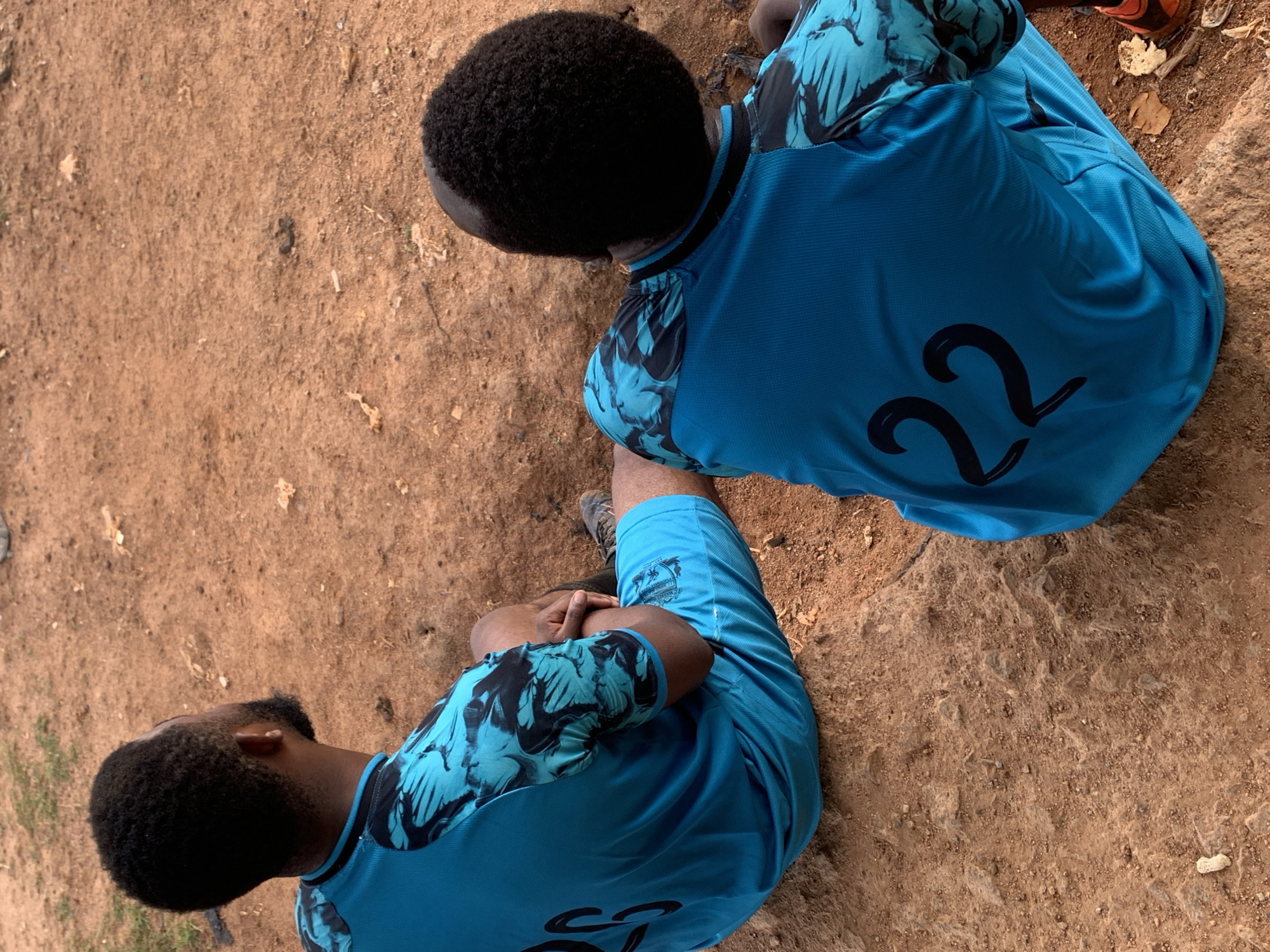 Two individuals, viewed from behind, are seated on a reddish-brown, dusty ground. Both are wearing bright blue athletic shirts with black and turquoise patterned sleeves. The person on the left has "25" printed in black on the back of their shirt, while the person on the right has "22" printed in black. Their legs are bent, and they are both wearing athletic shoes. The ground is uneven and scattered with small stones and dry leaves. The setting appears to be an outdoor area, possibly a sports field or park, in Bukuru, Nigeria, characterized by its reddish soil and sparse vegetation.