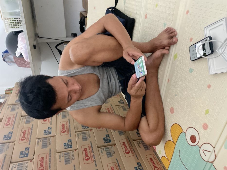 An adult male of Asian ethnicity is depicted sitting cross-legged on a patterned floor mat, engrossed with a white smartphone held horizontally in both hands. He is wearing a grey sleeveless shirt and dark blue shorts, and is barefoot. His gaze is directed downwards at the smartphone screen, which displays a grid of application icons.
To the man's lower right, a dark blue or black smartphone lies face-up on an open white rectangular box, likely its retail packaging. A white charging cable is connected to this phone, coiled next to it. The floor mat features a light background with subtle patterns and a larger, colorful cartoon-like design (partially visible, depicting a blue and yellow element with the number "7") in the bottom left corner of the frame.
The background on the left is dominated by a tall stack of beige cardboard boxes, branded "Omela KRIMER KENTAL MANIS" and indicating "2 x 500G BAG" along with the code "1251779". This suggests a storage or retail environment. On the right, a white shelf or table holds a black motorcycle helmet, a light pink item (possibly a bag or textile), and a brown patterned bag. A white plastic basket is also visible on the shelf. The scene appears to be an indoor setting within Kayen, Indonesia.