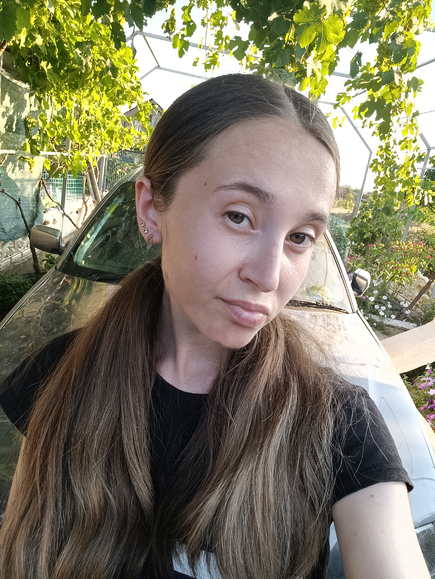 This is a selfie taken outdoors, likely in a rural or suburban area, with a woman in the foreground. She has long, light brown hair pulled back into a ponytail. She is wearing a black t-shirt and has piercings in her left ear. Behind her, a silver car is visible, with foliage and a trellis overhead. The background appears to be a yard or garden with plants and flowers. The lighting suggests it is daytime, possibly late afternoon. The location is Vypasne, Ukraine.