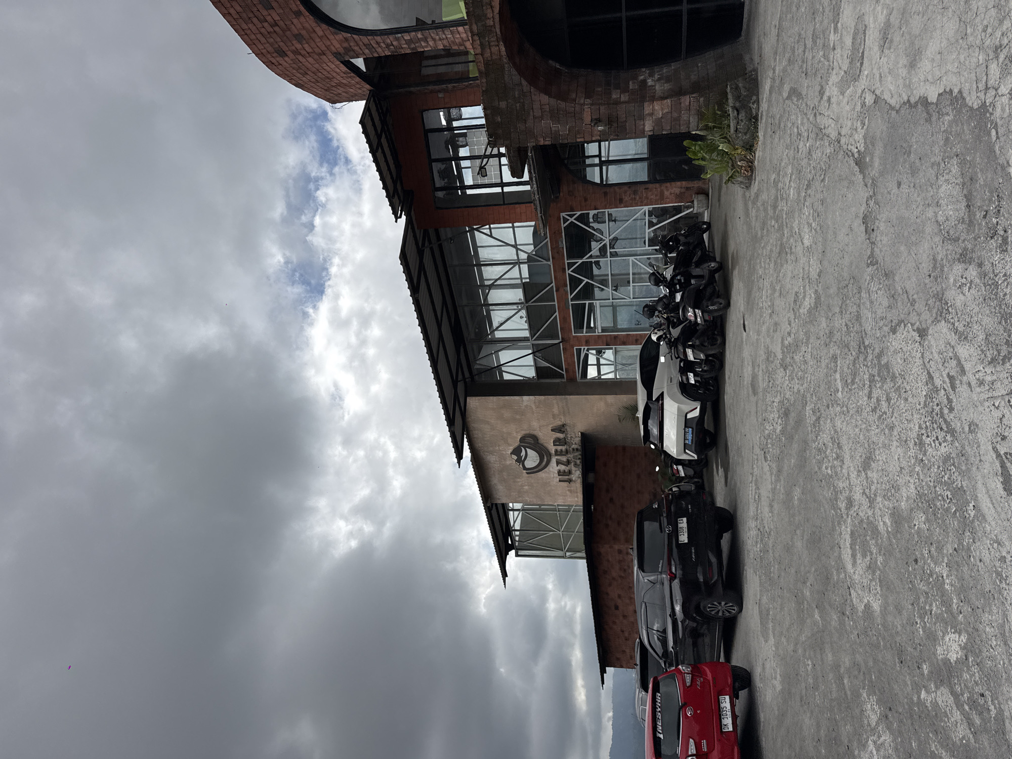 The image shows the exterior of Jezera Cafe in Kintamani, Indonesia, on a cloudy day. The cafe building has a modern design with large glass windows and brick accents. Several vehicles, including cars and motorcycles, are parked in the paved area in front of the cafe. The scene appears to be a public space, likely a tourist attraction, given the presence of multiple vehicles and the cafe's prominent signage. The surrounding environment is somewhat obscured by the building and the cloudy sky, but hints of mountainous terrain and vegetation can be seen in the distance.