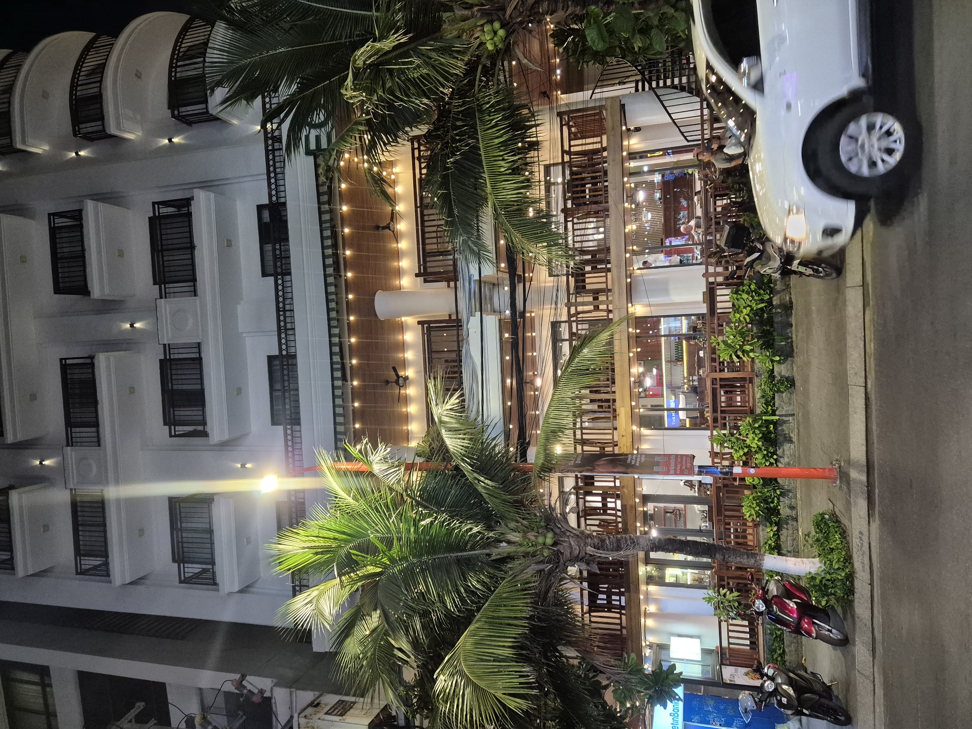 The image depicts a multistory building with a modern facade, likely a hotel or apartment complex, at night in Đà Nẵng, Vietnam. The building is illuminated by streetlights and strings of warm-toned fairy lights, creating a festive atmosphere. A large palm tree with coconuts stands prominently in the foreground, its leaves lush and green.
On the ground floor, there appears to be a restaurant or cafe with open-air seating, evidenced by tables and chairs visible through the windows. Several motorcycles are parked along the sidewalk, suggesting a common mode of transportation in the area. A white SUV is driving in the street in the foreground, its headlights on.
Several people are visible in the image: two individuals are seated at a table in the restaurant area, and another person is standing near the entrance. The overall scene suggests a lively urban environment, with elements of both commercial activity and residential living. The architecture and vegetation are consistent with a tropical coastal city like Đà Nẵng.