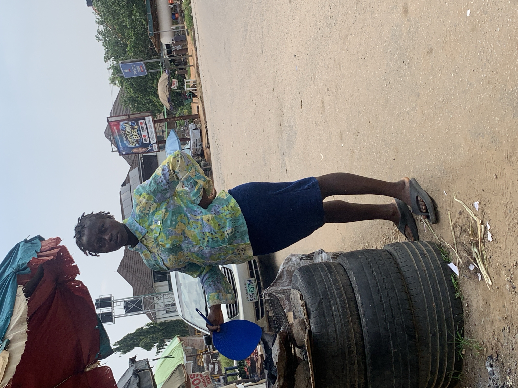 A person stands in a street scene, holding a blue fan in their left hand. They are wearing a colorful, patterned shirt and a dark blue skirt. The ground is a sandy, dusty surface, and to their right are three stacked tires. In the background, there are some stalls or makeshift shops with tarpaulin coverings, a white vehicle with a Nigerian license plate (RSH-637MW), and various signs. One sign is a blue street sign with "BWARI" and "ONION" visible. Another sign is a large advertisement with text including "SUPERNATURAL EDGE." The sky is overcast, suggesting it could be a cloudy day. The overall impression is of an outdoor market or street-side business in an urban or semi-urban area in Abuja, Nigeria. The person's gaze is directed towards the camera, and their expression appears neutral. The scene seems to be during daylight hours, but the lighting does not suggest bright sun.