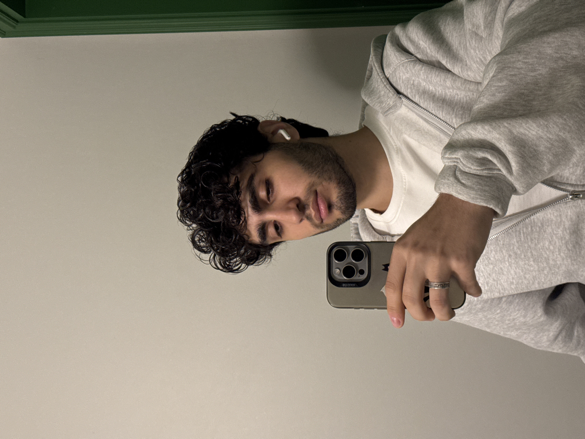 A man with dark curly hair is taking a selfie. He is wearing a white t-shirt and a gray zip-up hoodie. He is holding a gray iPhone in his right hand. He is wearing a silver ring on his right ring finger. He has an earbud in his left ear. The background is a plain wall with a green trim. The image provides no information about Yerevan, Armenia.
