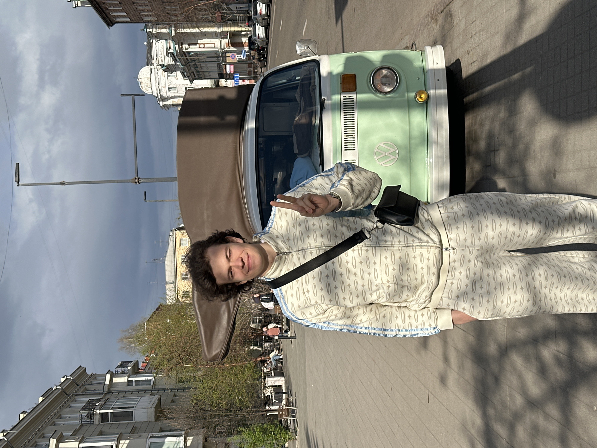 A young man stands on a sidewalk in Krasnoyarsk, Russia, giving a peace sign. He is wearing a patterned white tracksuit with blue trim and a black crossbody bag. He is standing next to a vintage green and white Volkswagen van. In the background, there are city buildings, a sky with some clouds, street lights, trees, and people seated at tables, likely in a cafe or restaurant area. The architecture visible includes a building with a dome.