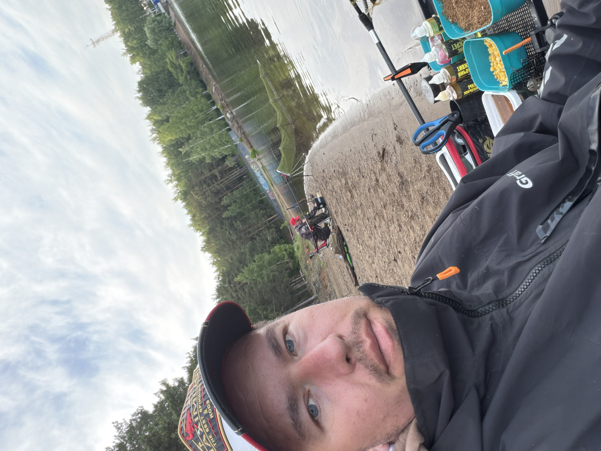 A man takes a selfie while fishing in Chaikovsky, Russia. He is positioned in the foreground, wearing a black jacket with an orange zipper pull (displaying "Graff" text upside down) and a baseball cap partially visible with "THE BEST SPINNING RODS" text. He looks towards the camera with a neutral expression, revealing light blue eyes and light stubble.
The setting is a tranquil fishing spot by a body of water, likely a river or lake, with a sandy/gravelly bank. Fishing rods are set up, some visible against the water. A fishing station is equipped with two blue containers holding bait: one with granular ground bait and another with yellow corn kernels. Several bottles of liquid attractants or additives are also present, featuring Cyrillic text such as "HONKI," "SWEET BOUQUET," "KRIL," and "ROACH." A pair of blue-handled scissors is attached to the station. A green keepnet is partially submerged in the water near the bank.
In the background, a dense line of evergreen trees reflects in the calm water on the opposite bank. The sky is cloudy, suggesting an overcast day. A tall, lattice-like tower is visible on the distant horizon above the treetops. The scene captures a peaceful daytime fishing excursion.