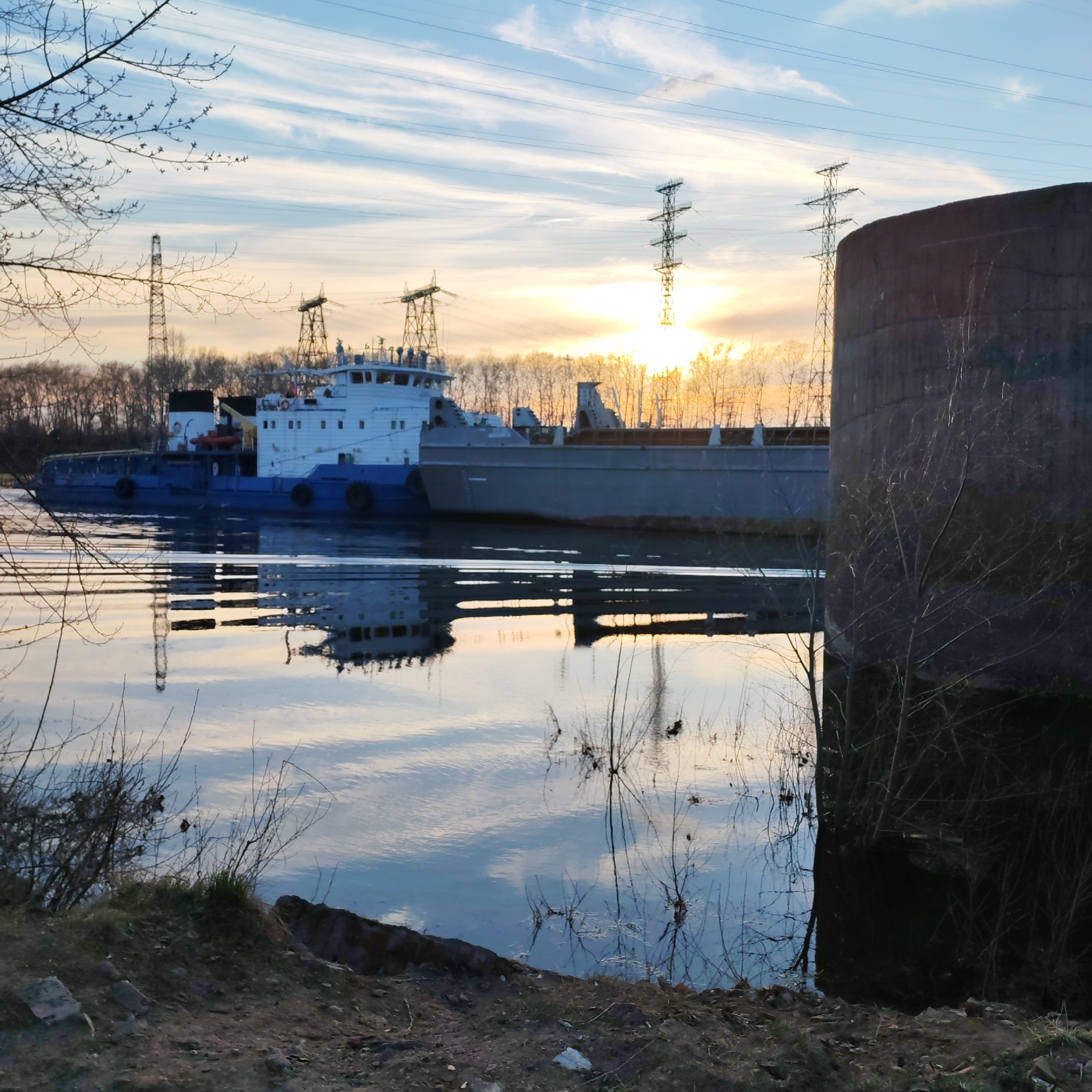 The image shows a tugboat with an attached barge on the Kama River, possibly in the vicinity of Chaikovsky, Russia. The sky is visible with a sunset, and there are power lines running in the background. A concrete structure is to the right of the river, with sparse foliage in the foreground along the river bank. The image is taken from a low angle, emphasizing the water's surface and reflections.