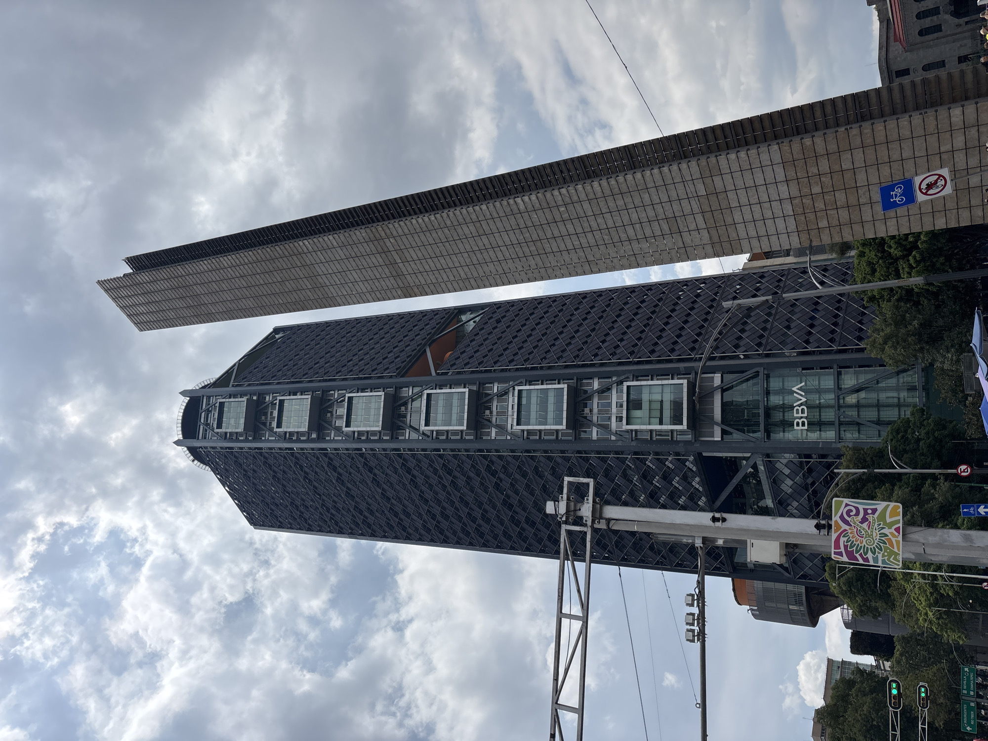 The image captures a street-level view of the Torre BBVA, a distinctive skyscraper in Mexico City, Mexico, characterized by its dark, latticed facade and illuminated glass boxes. To the right of the Torre BBVA, the Torrel T.A.L. also stands, distinguished by its slender, beige, and rectangular facade. The sky above is partially cloudy, with patches of blue visible. In the foreground, a traffic light with green signals is visible, along with street signs indicating directions to Insurgentes and Sevilla. A colorful, circular mural-like sign is affixed to a lamppost, and a blue sign with a bicycle symbol and a red circular sign with a bicycle crossed out are also present. Trees and other buildings are visible in the background, suggesting an urban environment. No people are clearly discernible in the frame.