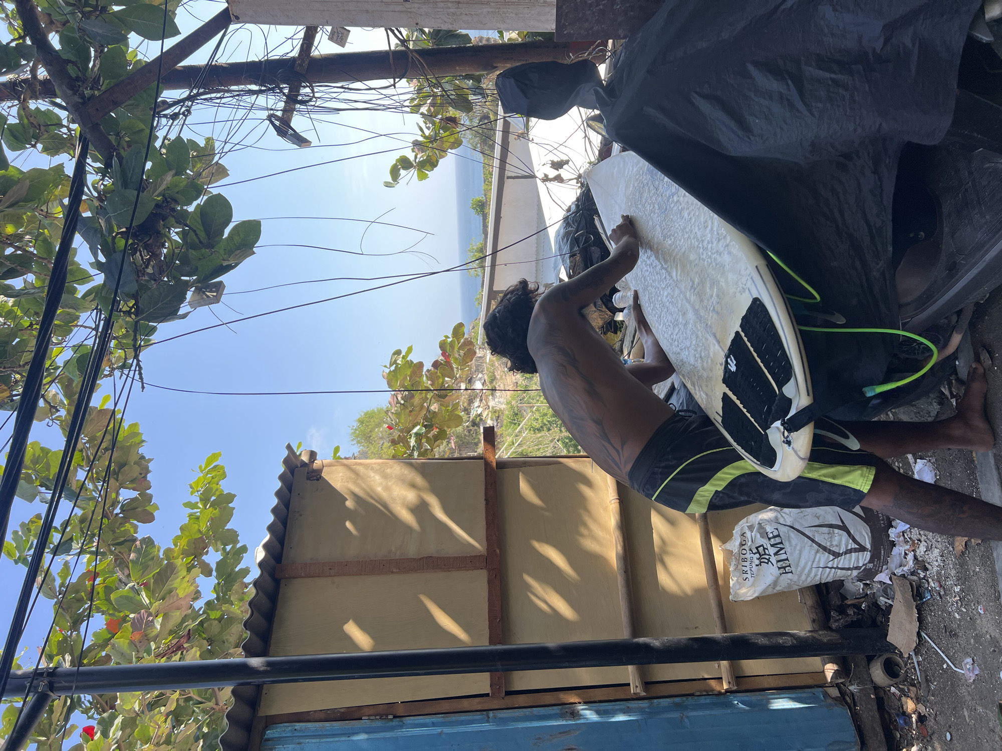 A man with extensive tattoos on his back and arms is waxing a surfboard. He is wearing black and green swim trunks. The surfboard is light-colored with black traction pads and neon green leash. He is positioned outdoors, in what appears to be a cluttered, utilitarian area, possibly a backyard or workshop, with a view of the ocean in the distance. A corrugated metal roof and wooden wall of a structure are visible behind him. Wires and branches of a tree hang overhead, casting shadows. A large bag of "SRIBOGA TEPUNG TERIGU HIME" flour is leaning against the wall. Debris is scattered on the ground. The scene suggests a casual, possibly tropical setting in Kuta Selatan, Indonesia, where a surfer is preparing his equipment.