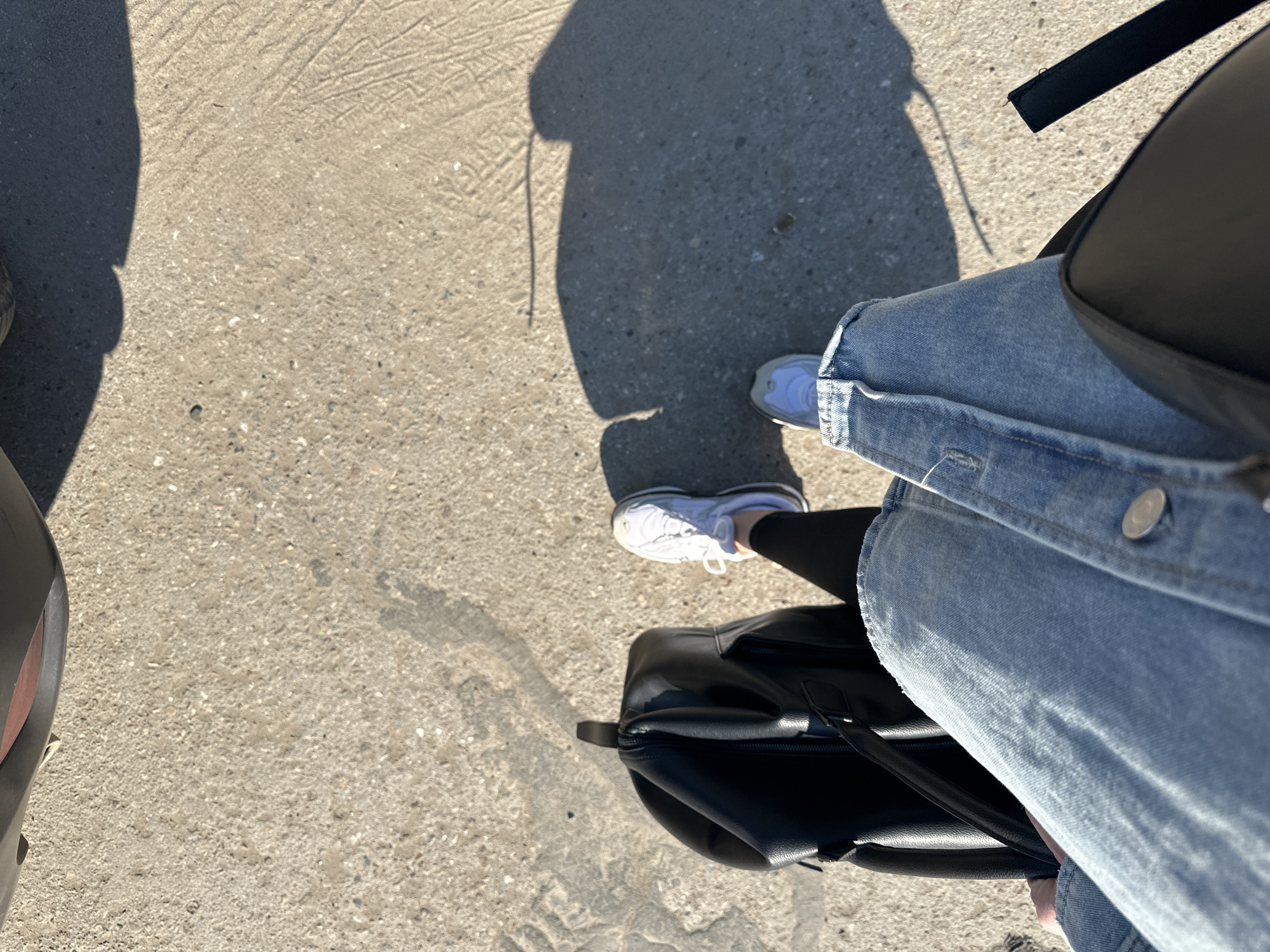 The image depicts a first-person view from above, looking down at a person standing on what appears to be a concrete surface. The person is wearing white sneakers, blue jeans and has a black bag resting against their leg. A shadow of the person is visible on the concrete, suggesting outdoor lighting. Part of what appears to be a car or other vehicle is visible on the left side of the image. No specific details indicate the location is in Chaikovsky, Russia.