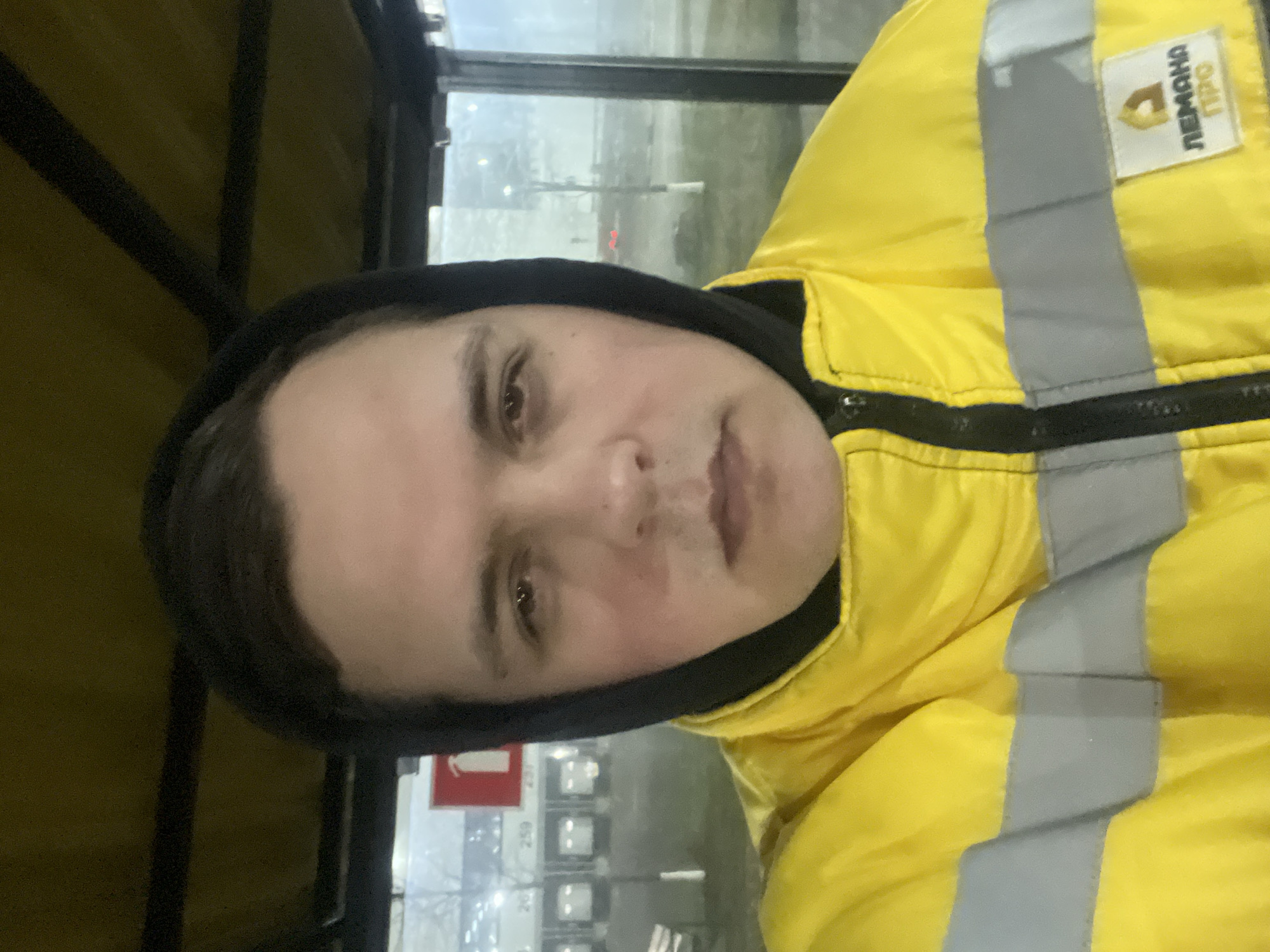 A medium shot depicts a person with fair skin, dark hair, and light-colored eyes wearing a yellow jacket with reflective grey stripes and a black hooded sweatshirt. The person's face is centrally framed, displaying a neutral expression. The jacket features a small white patch with lettering in Cyrillic script (likely "Лемана ПРО").
In the blurred background, there appears to be an urban environment, possibly a bus station or covered waiting area, with signage in Russian, including a red and white sign. Further in the distance, light-colored structures or buildings are visible through the window.
Given the Cyrillic text and urban setting, the image is likely taken in a Russian-speaking area; however, the location cannot be confirmed to be Lupanovo, Russia based on the available image data alone. More specific details would be needed to pinpoint the exact location.