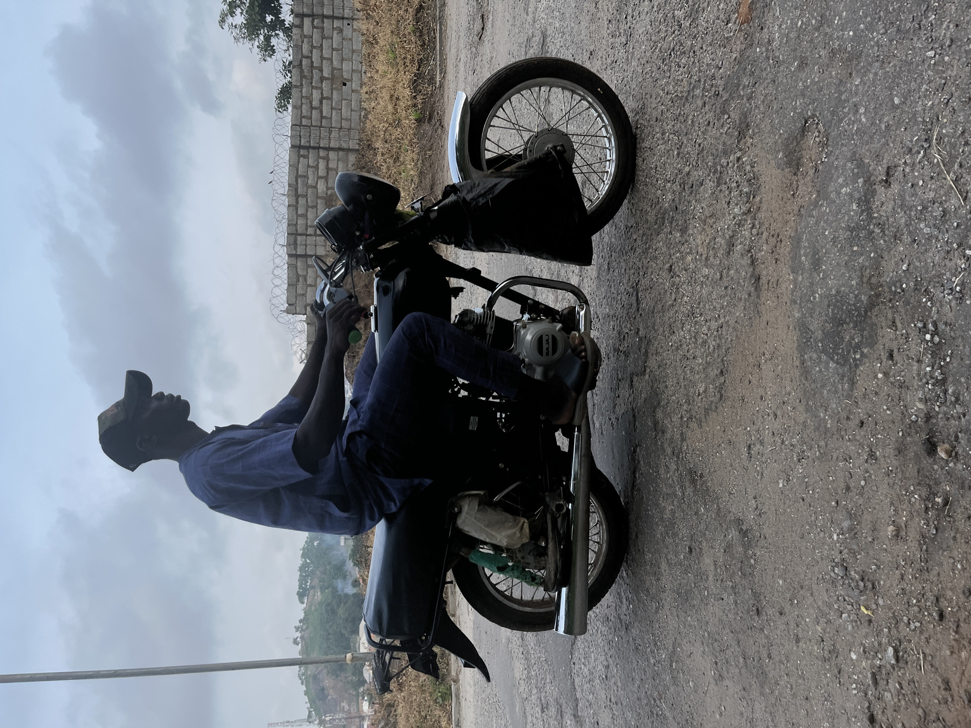 A person wearing a blue shirt, dark pants, and a baseball cap rides a black motorcycle on a gravel road. The person is positioned in the middle of the frame, facing to the right. The motorcycle has a chrome exhaust pipe and a black fender. In the background, there is a brick wall with barbed wire on top, dry grass, and a cloudy sky. The scene appears to be in an outdoor urban environment, possibly in Abuja, Nigeria, given the style of the motorcycle and the architecture of the wall.