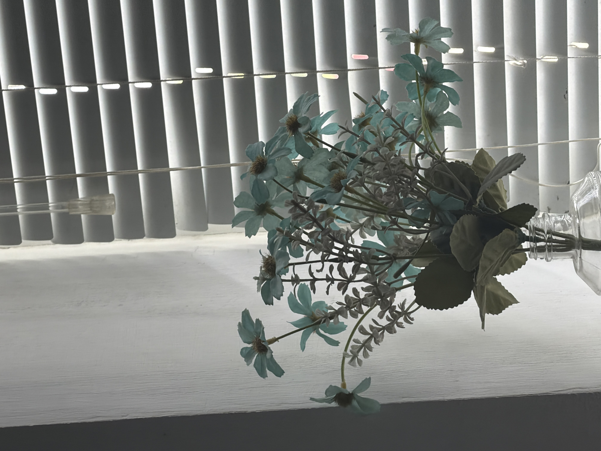 A close-up, eye-level shot focuses on a bouquet of artificial blue flowers in a clear glass vase. The vase is positioned on a surface, partially obscured by the lower part of the bouquet. The flowers, in various shades of blue and teal, are arranged with some green foliage and smaller, grayish-green filler flowers. The background is divided: on the left is a textured white wall, and on the right are white horizontal blinds with light filtering through them. A thin cord hangs vertically in front of the blinds. The lighting suggests it's daytime, with light coming from the right, casting subtle shadows and highlights on the flowers and blinds. There are no people or animals present.