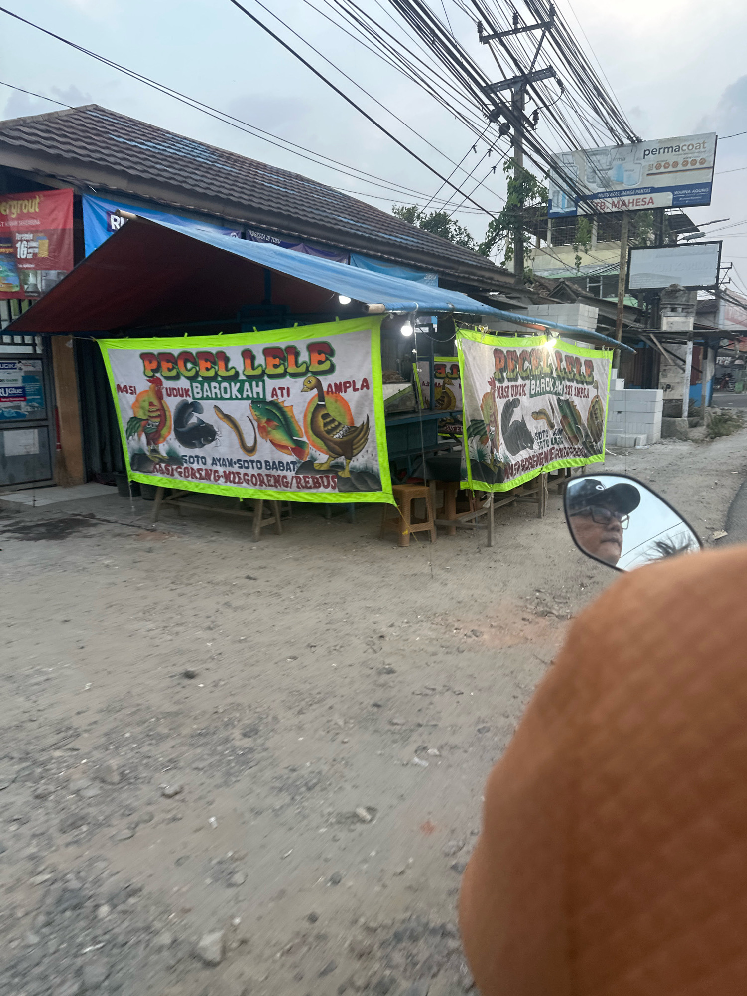 The image depicts a roadside food stall in Saruni, Indonesia, specializing in "Pecel Lele," a dish of fried catfish. The stall is adorned with a large banner featuring cartoonish illustrations of a chicken, cat, eel, fish, and duck, along with the text "PECEL LELE NASI UDUK BAROKAH ATI AMPLA SOTO AYAM-SOTO BABAT NASI GORENG-MIE GORENG/REBUS". The stall itself is covered by a blue and red awning, and wooden stools are visible. In the foreground, a motorcycle's side mirror reflects the image of a person wearing glasses and a baseball cap. The ground in front of the stall is unpaved, with visible dirt and gravel. Various electrical wires crisscross the sky above the stall, and a billboard advertising "PERMACOAT" and "MAHESA" is visible in the background.