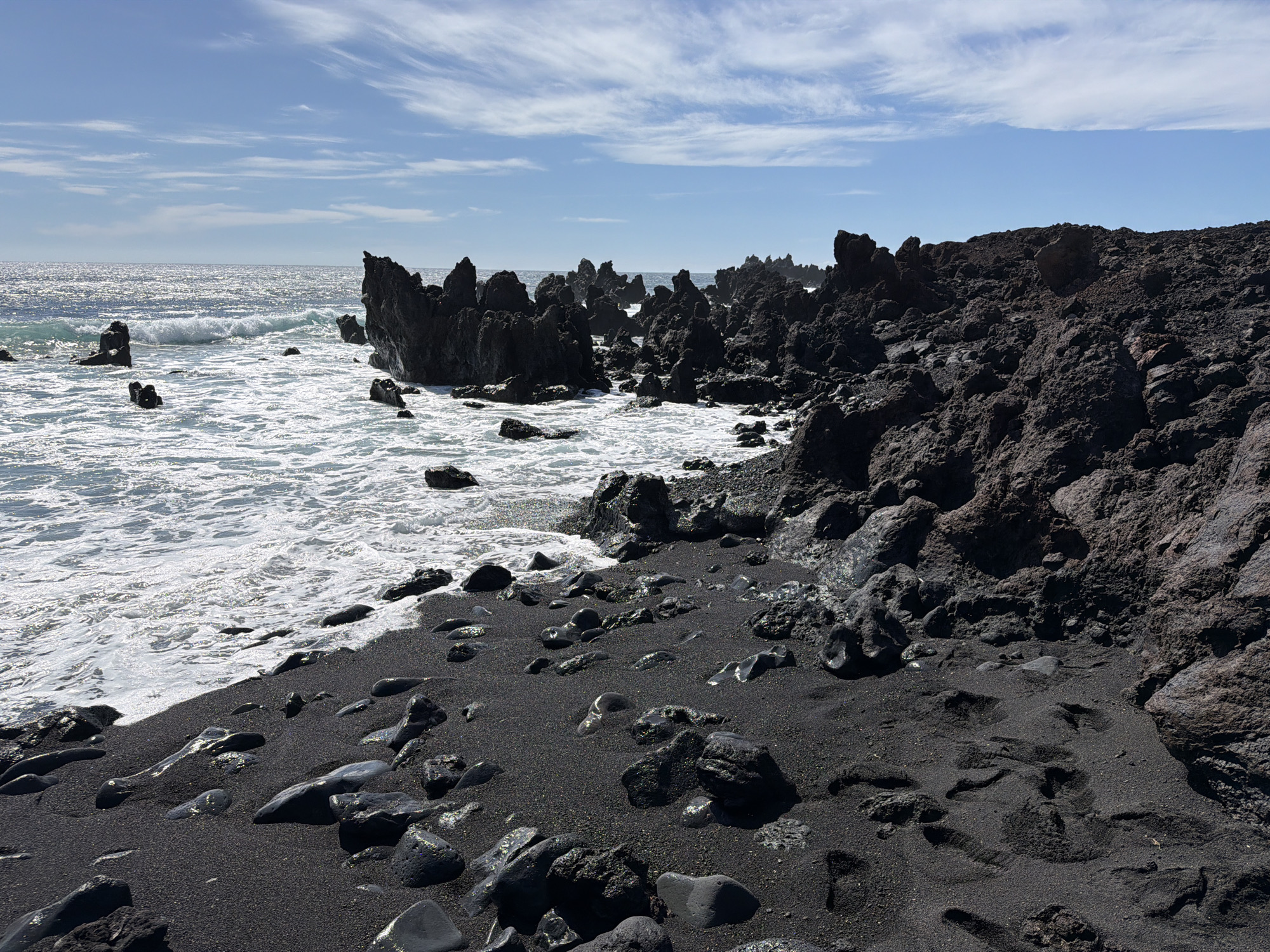 The image depicts a black volcanic sand beach in Yaiza, Spain. Sharp, black rock formations rise from the sand and extend into the ocean, where waves are visible. The sky is blue with wispy white clouds. The sand is dark and coarse, with larger, smooth black stones scattered across it. A rocky slope forms the edge of the beach, transitioning from the sand to a higher ground of volcanic rock. The ocean water is foamy where it meets the shore.