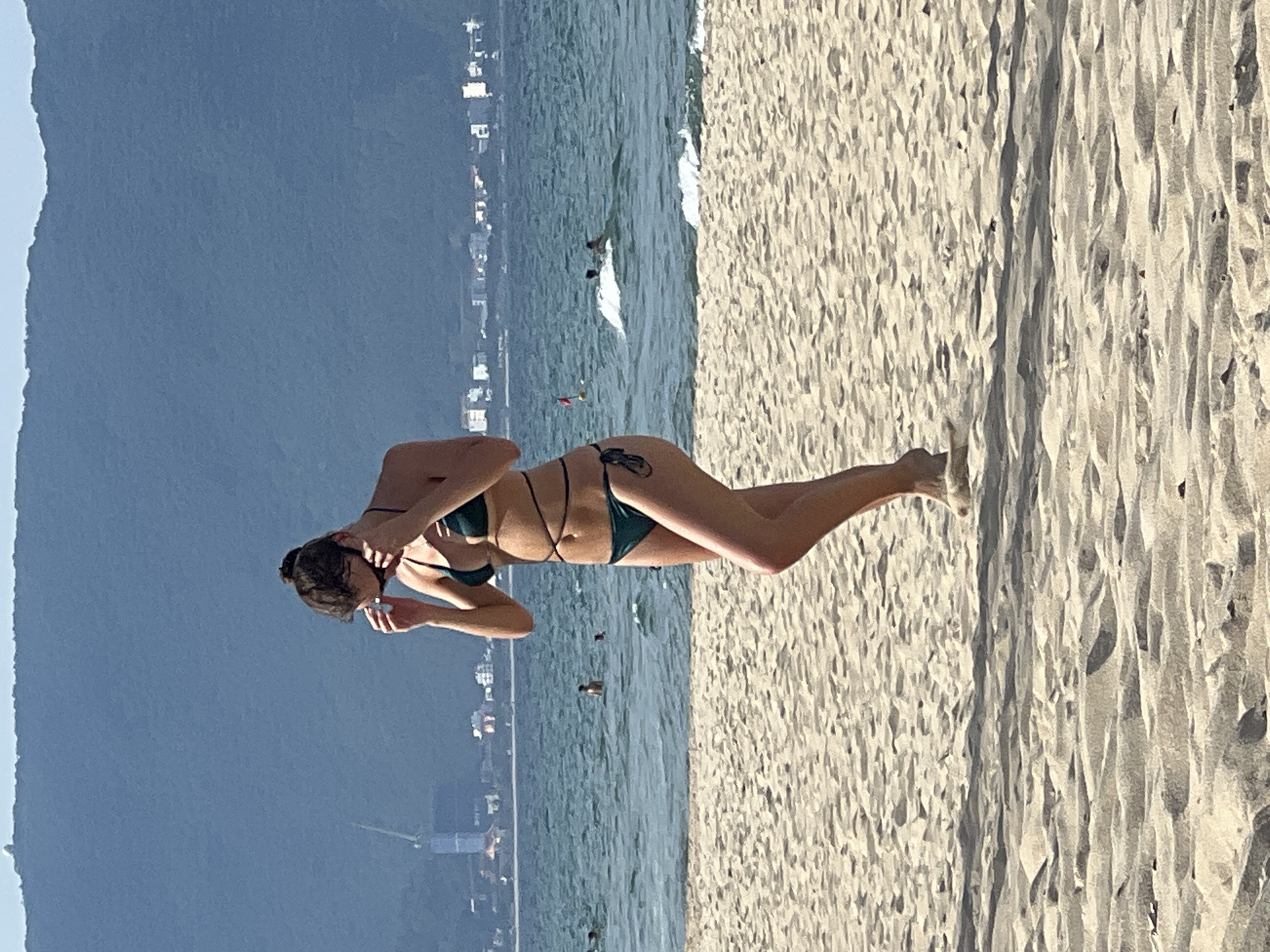 A young woman in a dark green bikini stands on a sandy beach in Đà Nẵng, Vietnam. She is looking down and adjusting her sunglasses, with her hands near her face. Her bikini has thin straps that tie around her waist. The ocean is visible behind her, with small waves and a few other people in the water. In the distance, there are buildings and a large, forested mountain. The sand is light brown and textured, with shadows cast by the uneven surface. The sky is clear and blue.