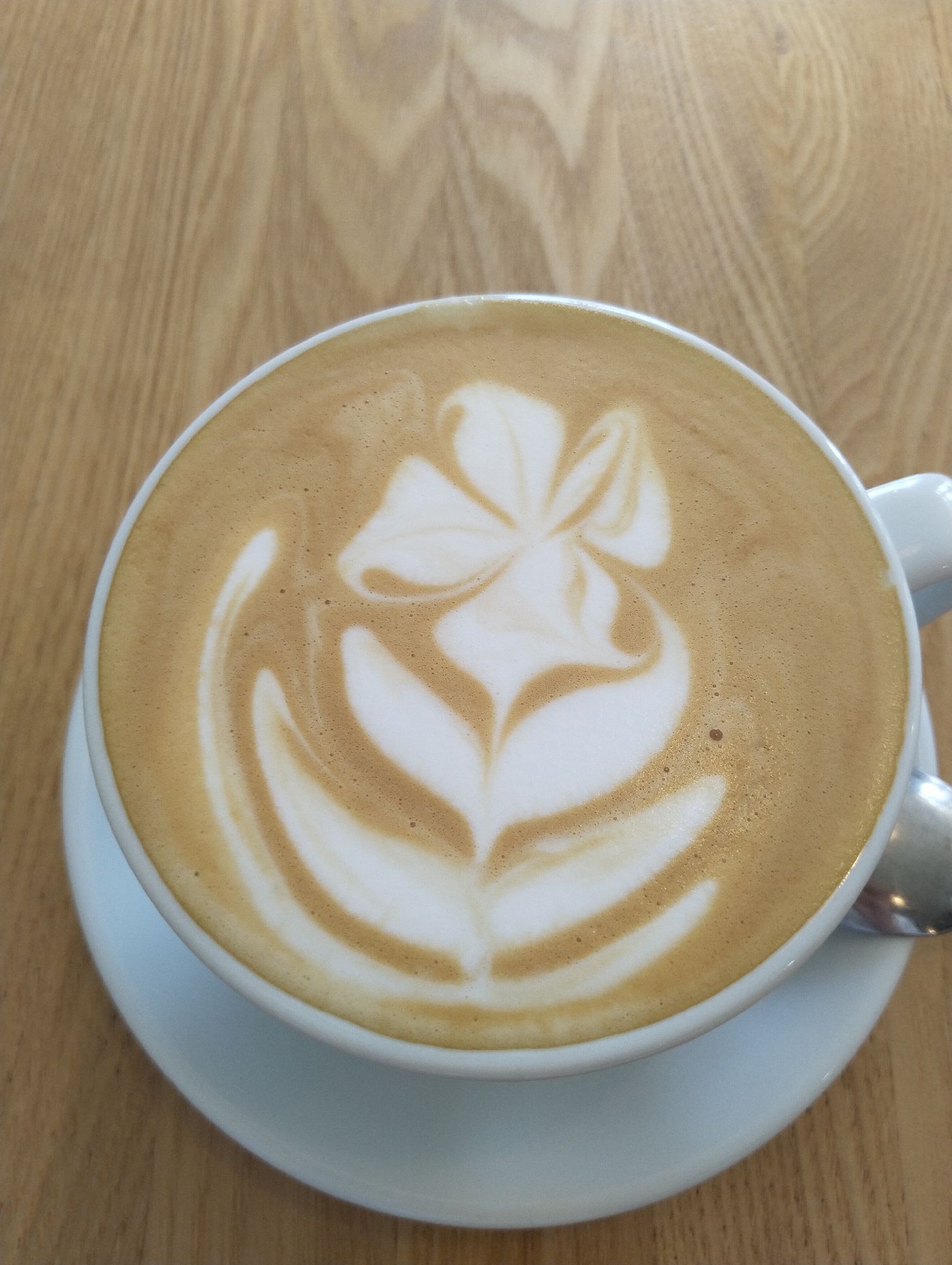 A top-down shot shows a cup of coffee on a saucer, placed on a light wooden table. The coffee features latte art in the form of a stylized flower. A spoon rests partially on the saucer, near the cup's handle. The environment suggests a cafe or similar establishment. There is no indication of location or people.