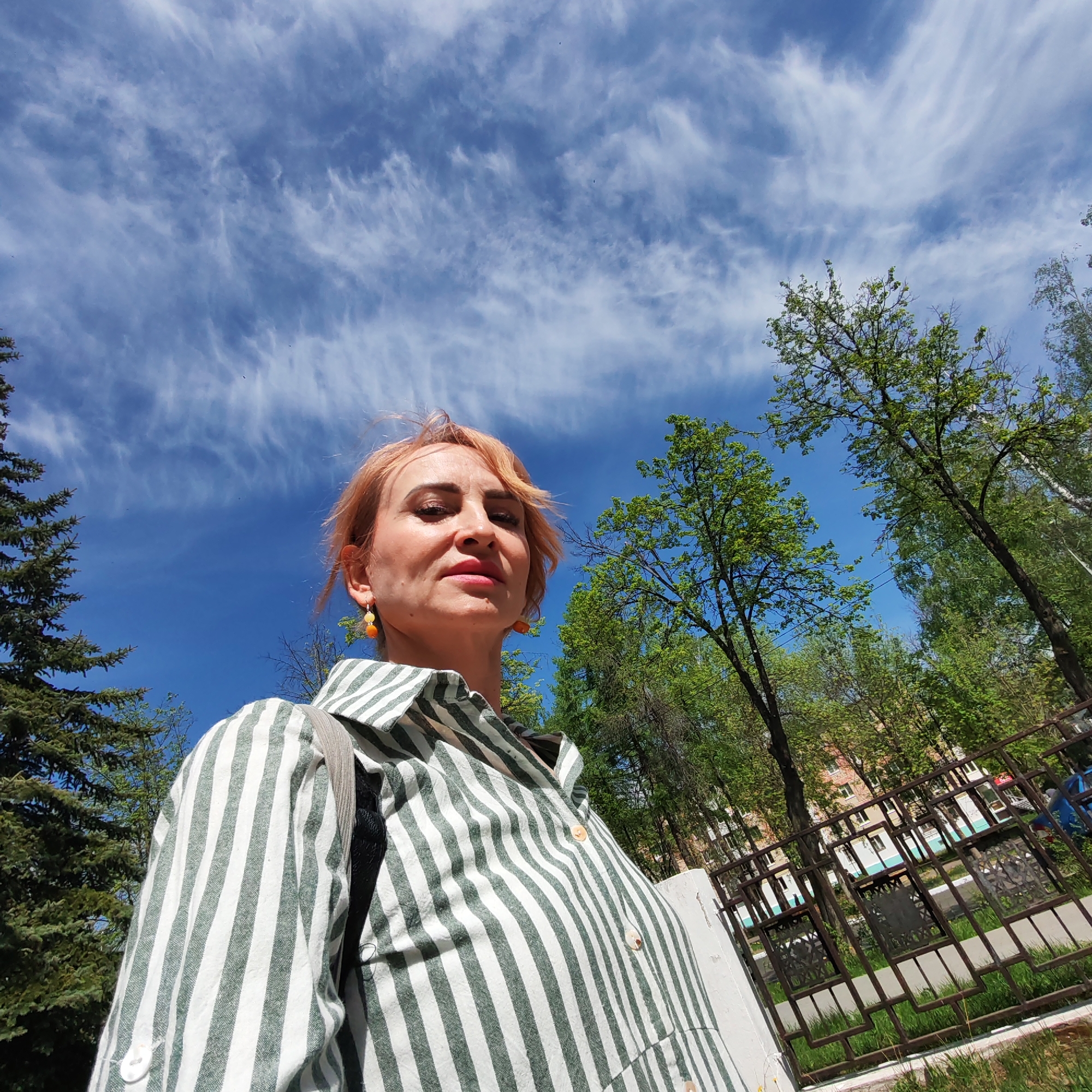 A woman with short, dyed red hair, wearing a striped green and white shirt and orange earrings, stands outdoors in Chaikovsky, Russia. She looks upwards towards the camera. Behind her is a bright blue sky with streaky white clouds. To the left is a tall evergreen tree. To the right, deciduous trees are visible, along with a wrought iron fence, a sidewalk, a lawn, and a multi-story building in the background.