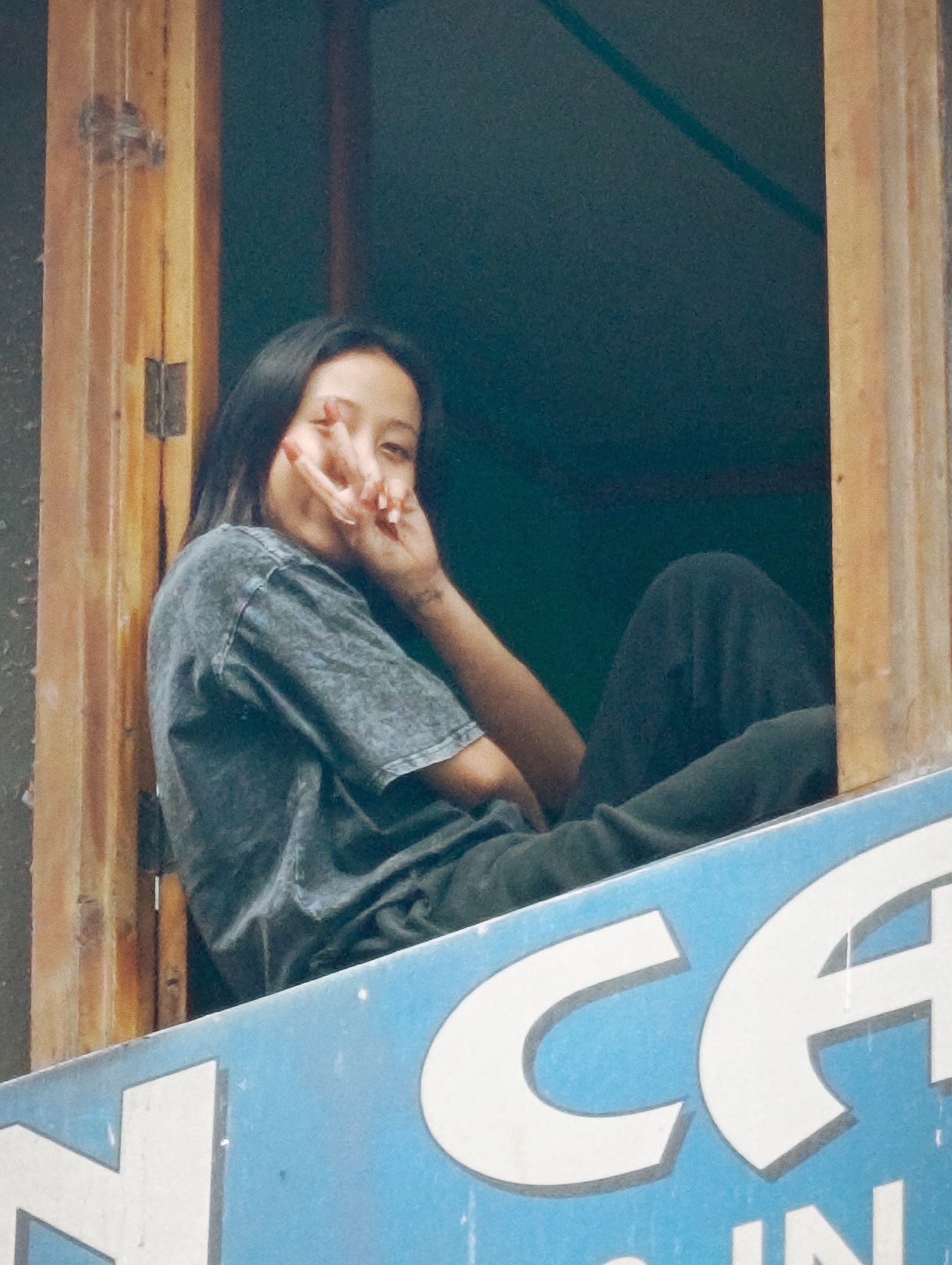 A young woman with dark hair is seated in a window frame, looking towards the viewer. She is wearing a dark gray t-shirt with a mottled pattern and dark pants. Her right hand is raised, with her index and middle fingers extended in a peace sign gesture. A tattoo is visible on her left forearm. The window frame appears to be made of weathered wood. In the foreground, a portion of a blue sign with white lettering is visible, displaying the letters "CA" and "N". The background is dark and out of focus. This scene is located in Vashisht, India.