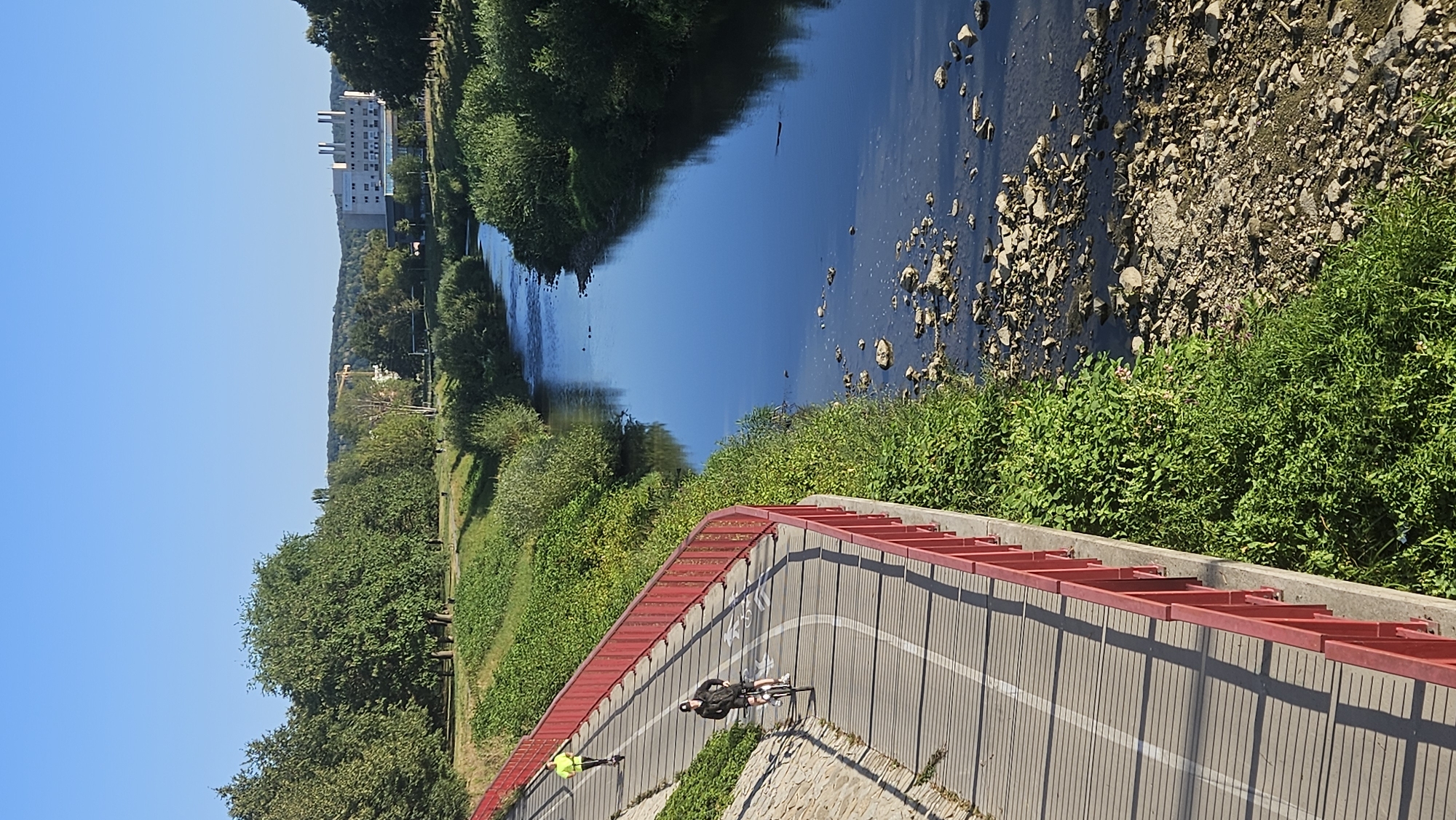 The image depicts a paved pathway for cycling and rollerblading, bordered by a red railing, running alongside a river in Prešov, Slovakia. A person is rollerblading in the distance, wearing a yellow jacket, and a person on a bicycle is closer to the foreground, wearing a dark jacket and a cap. The river has a rocky bed visible in parts and is surrounded by lush green vegetation and trees. In the background, across the river, a large, modern building with multiple ventilation shafts on its roof is visible, along with more trees and hills. The sky is clear and blue, suggesting a sunny day. The pathway is marked with white lines and symbols indicating bicycle lanes.