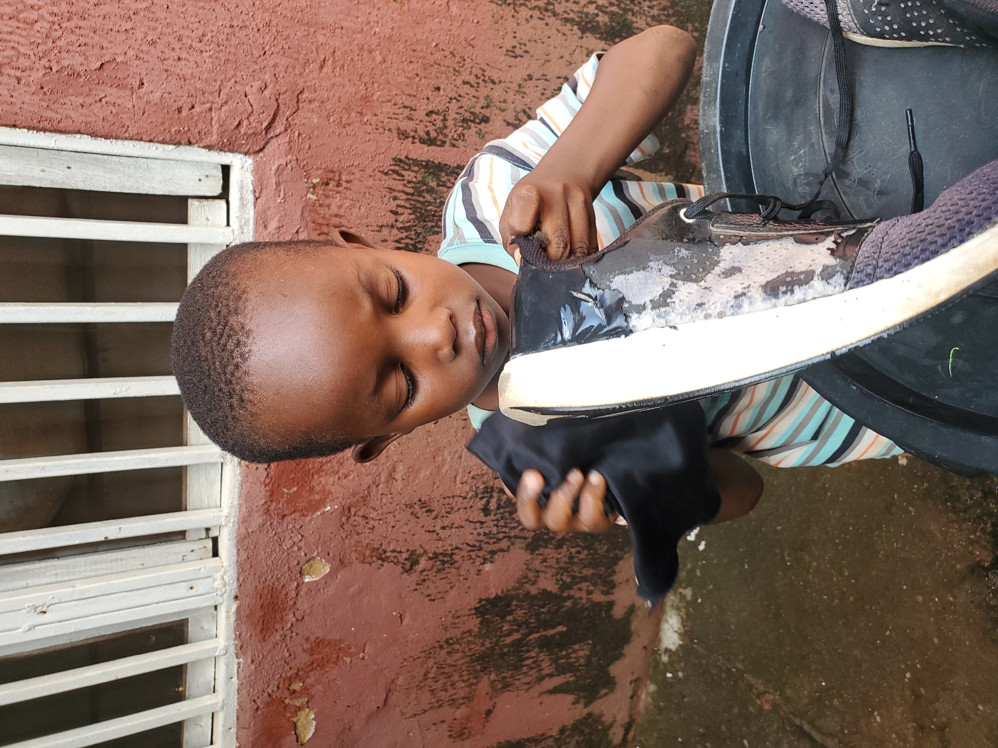 A young boy with dark skin and short hair, wearing a horizontally striped t-shirt, is diligently cleaning a black and white sneaker with a black cloth. He is positioned in front of a reddish-brown wall with peeling paint and a window with white vertical bars. The sneaker, which is wet and dirty, rests in a black tub. The ground is wet, suggesting it may have recently rained or that water is being used for cleaning. The scene appears to be outdoors, possibly in a residential area in Jos, Nigeria.