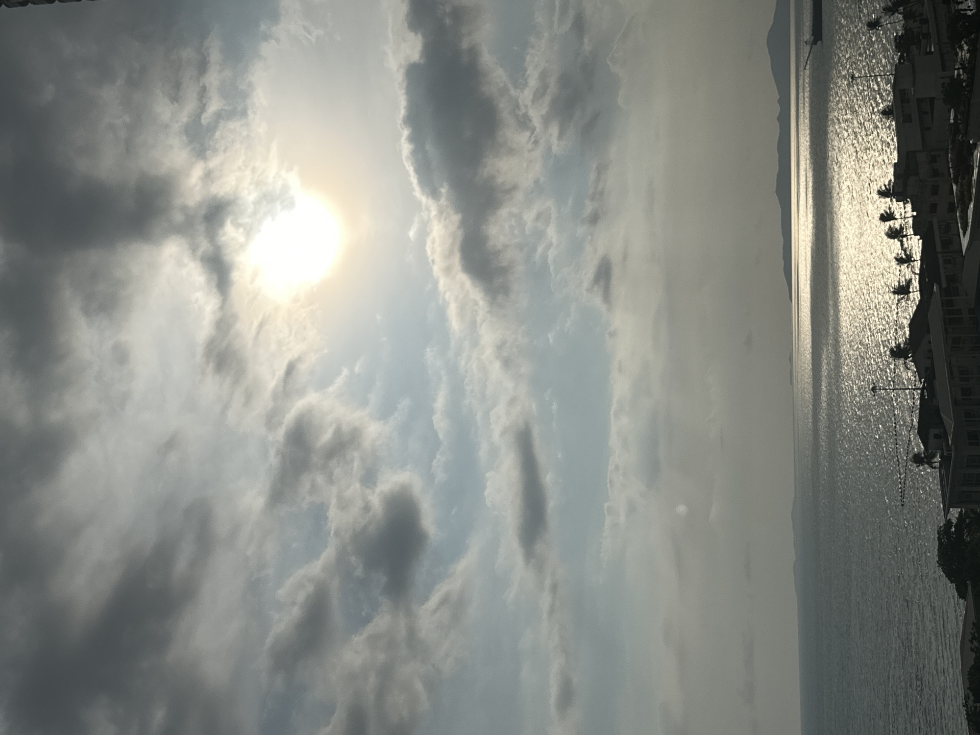 The image depicts a waterscape with the sun obscured by clouds in the sky. The water reflects the sunlight, creating a shimmering effect. On the right side of the image, buildings and palm trees are visible along the coastline. Given the prompt context, the location is likely in Pattaya City, Thailand.