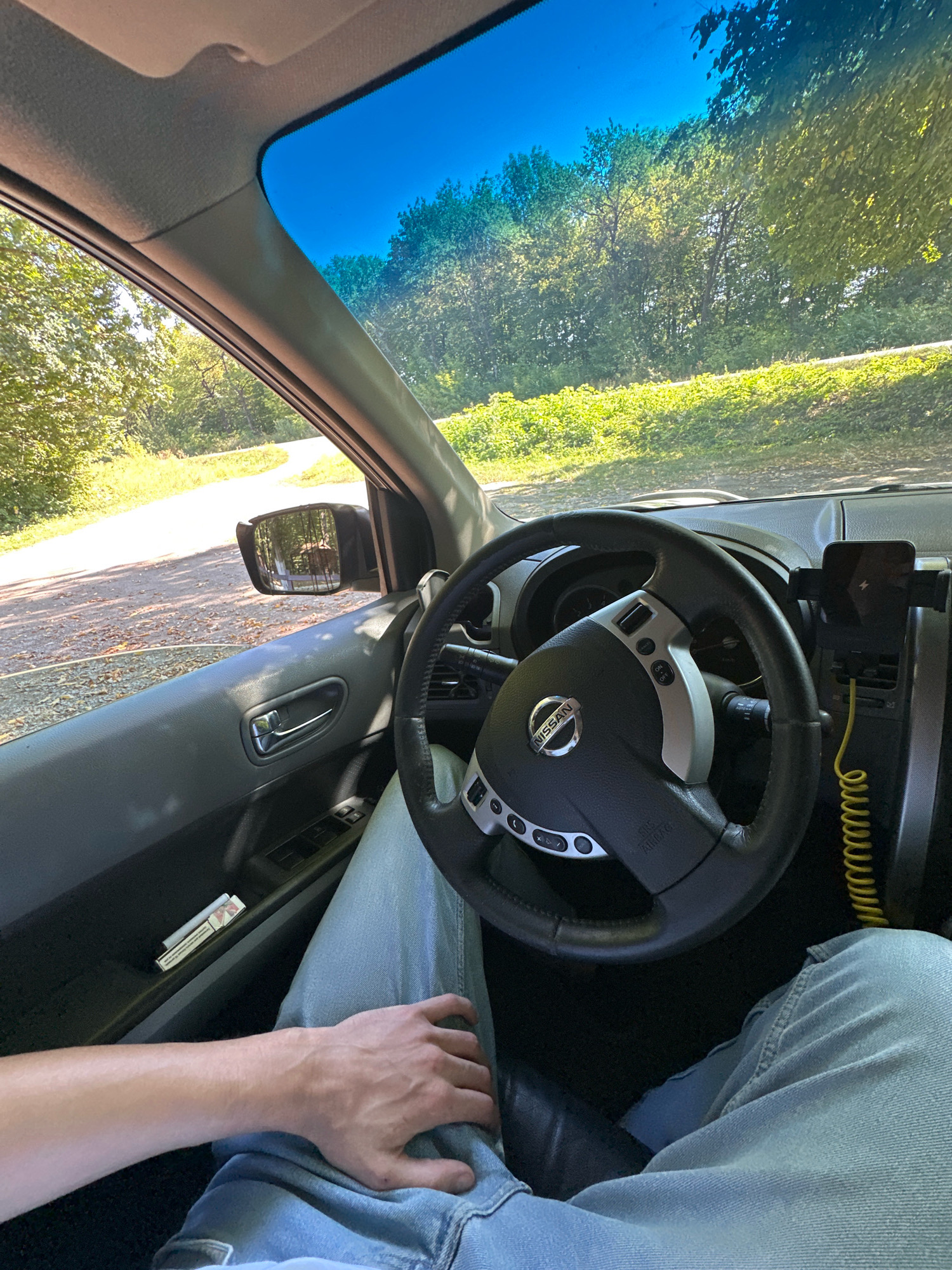A first-person perspective shot from inside a Nissan vehicle shows the steering wheel and dashboard, with the driver's left arm and leg visible. The driver is wearing blue jeans and their hand rests on their lap. A cigarette pack is in the door pocket. The car is on a dirt road bordered by lush green trees, suggesting a rural or wooded area. The sky is a bright, clear blue. The rearview mirror reflects a wooded scene. The location is identified as Shatava, Ukraine.