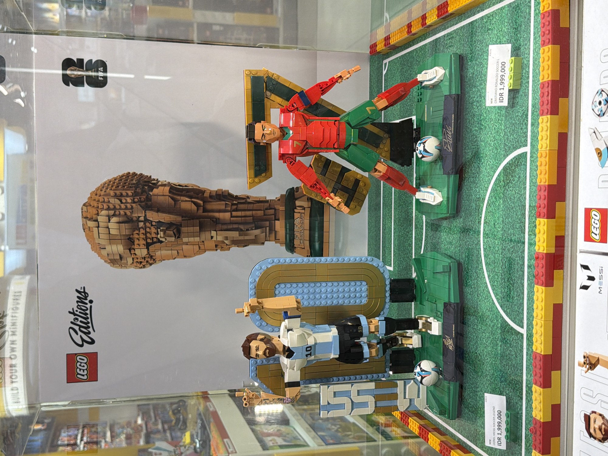 A LEGO display case in a store in Kuta, Indonesia features two iconic football player figurines, Lionel Messi and Cristiano Ronaldo, constructed entirely from LEGO bricks. The Messi figure, wearing the Argentina national team's blue and white striped jersey with the number 10, stands in front of a giant LEGO number "10" and a LEGO trophy. The Cristiano Ronaldo figure, in Portugal's red and green national team colors with the number 7, is positioned in front of a large LEGO number "7". Both figures are standing on a green LEGO football field, complete with white lines. A LEGO FIFA World Cup trophy is visible in the background. Price tags for both figures, reading "NEW LIONEL MESSI SOCCER LEGEND IDR 1,999,000" and "NEW CRISTIANO RONALDO SOCCER L IDR 1,999,000," are placed in front of their respective displays. The display is illuminated and encased in glass, suggesting it is part of a retail environment.
